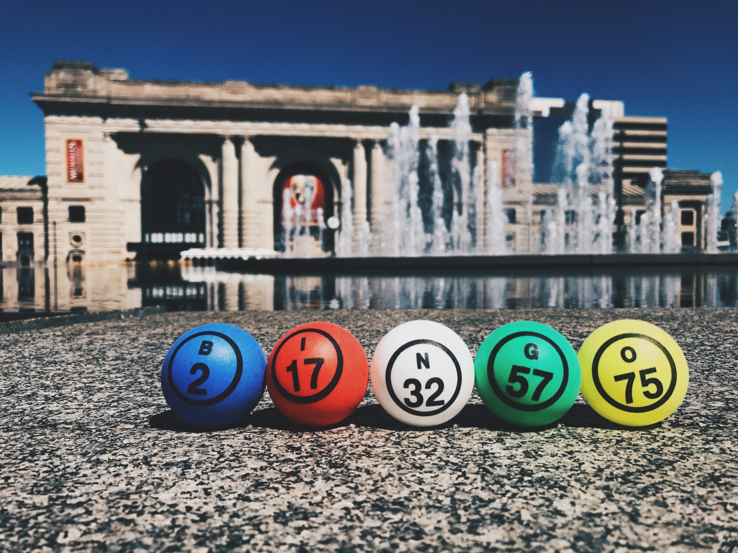 Five colorful bingo balls labeled B2, I17, N32, G57, and O75 are lined up on a ledge. Behind them is a historic building with columns and large windows, set against a clear blue sky with water fountains in the background.