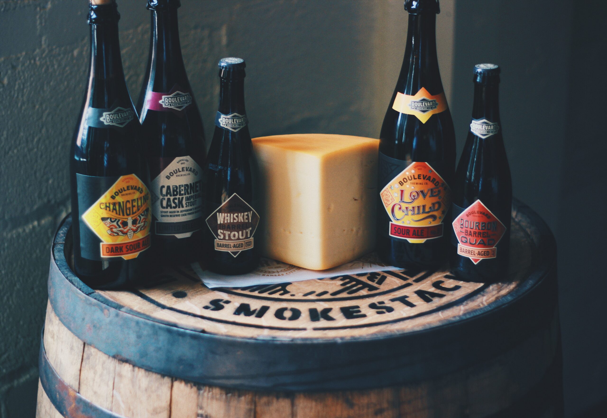 A selection of five Boulevard beer bottles is arranged around a large block of cheese on a wooden barrel labeled "Smokestack." The labels indicate different beer varieties, including "Whiskey Stout" and "Cabernet Cask." The setting is dimly lit.