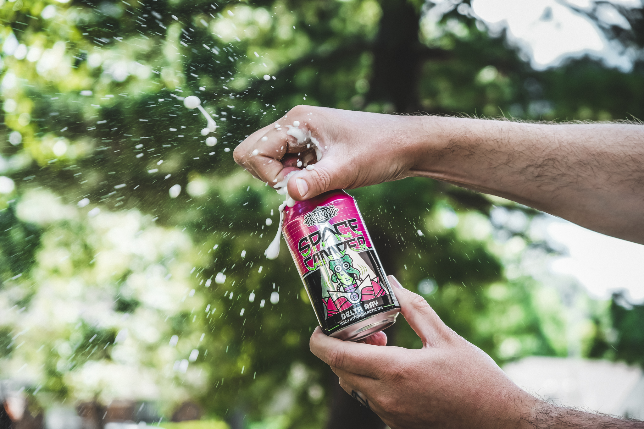 A person opens a can of "Space Zooider" beer, causing it to fizz and spray. The can features colorful graphics including space-themed artwork. The background shows trees and an out-of-focus park setting, suggesting an outdoor environment.