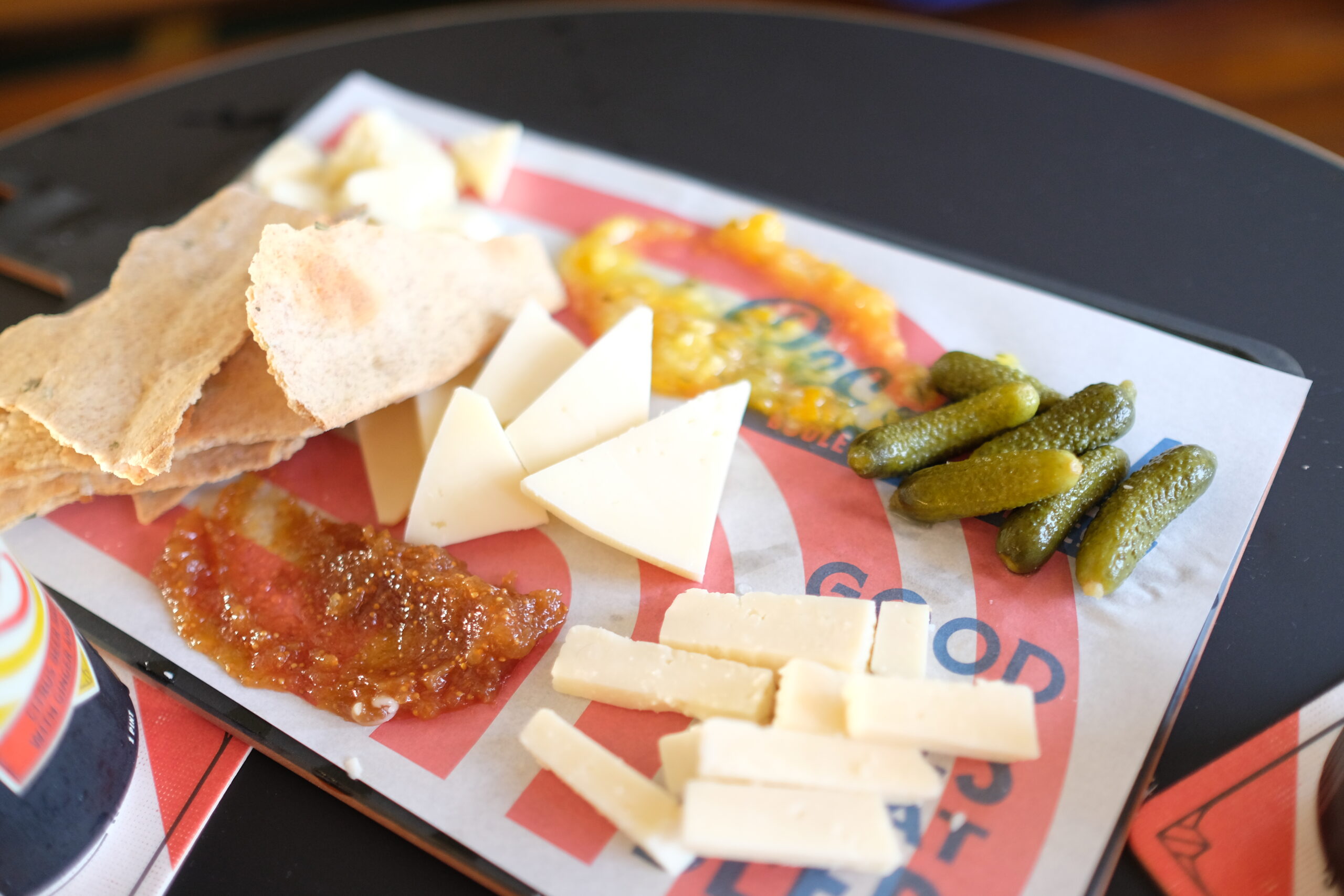 A tray with a variety of snacks including triangular and rectangular pieces of cheese, small pickles, crispy bread pieces, and two types of spreads or jams, one yellow and one red. The tray is placed on a black surface.