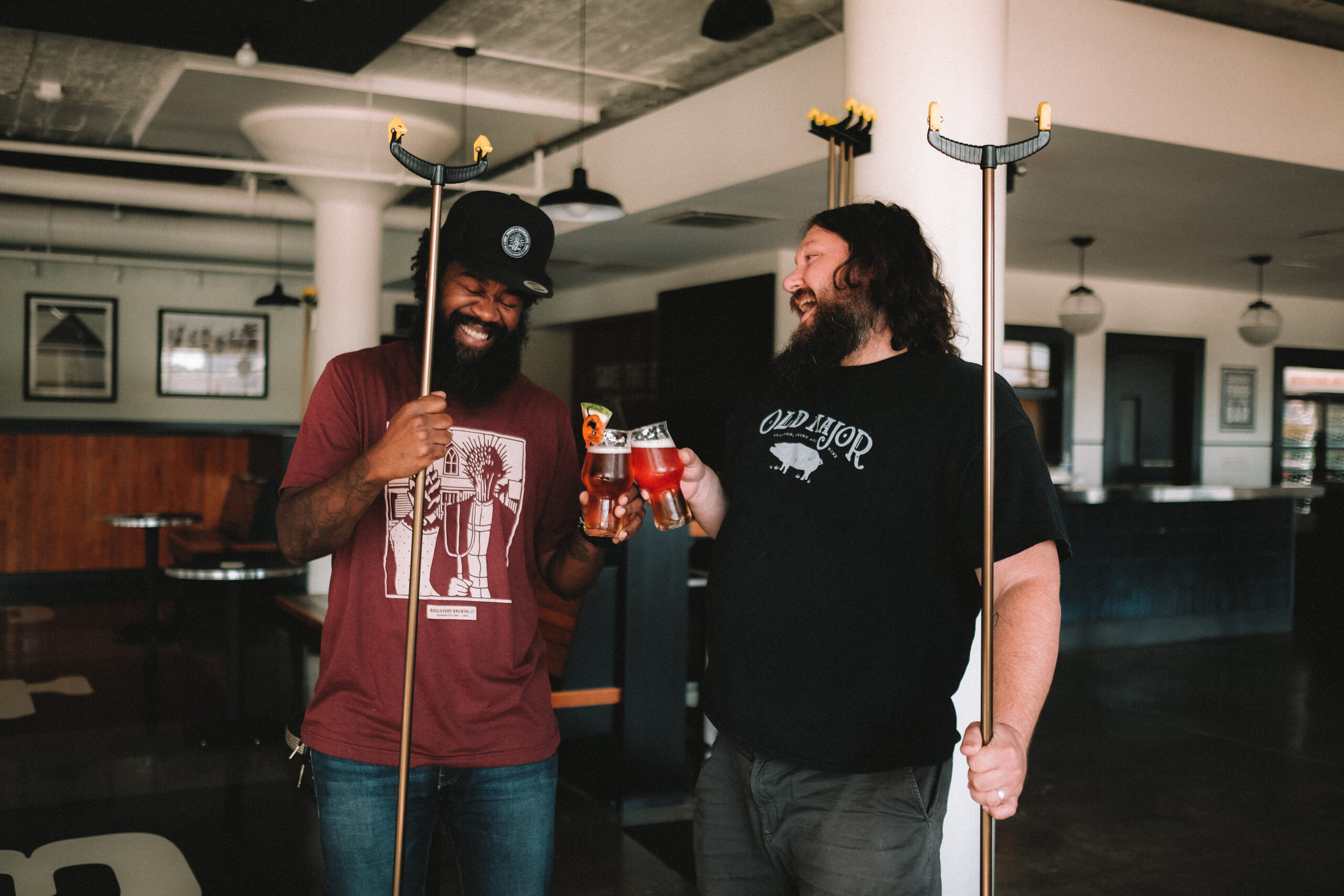 Two men in a bar, each holding a glass of beer and a shuffleboard cue, are smiling and clinking glasses. The man on the left wears a red shirt, and the man on the right wears a black shirt. Framed pictures hang on the walls in the background.
