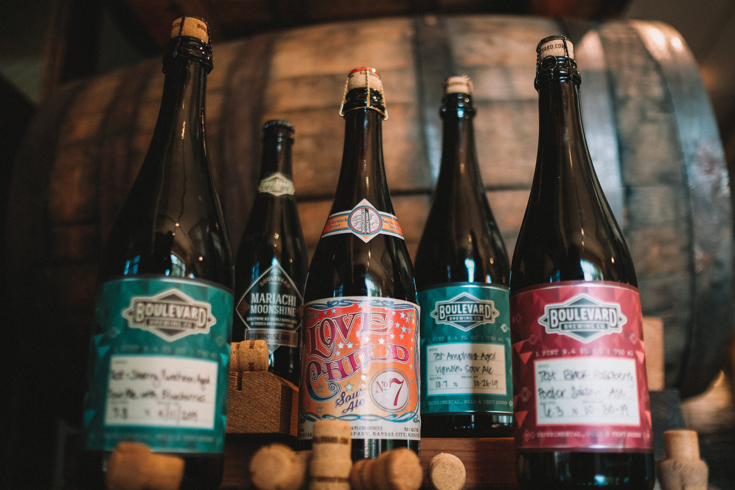 Five Boulevard beer bottles with colorful labels are displayed in front of a wooden barrel. Corks are scattered at the base of the bottles, which vary in design and color.