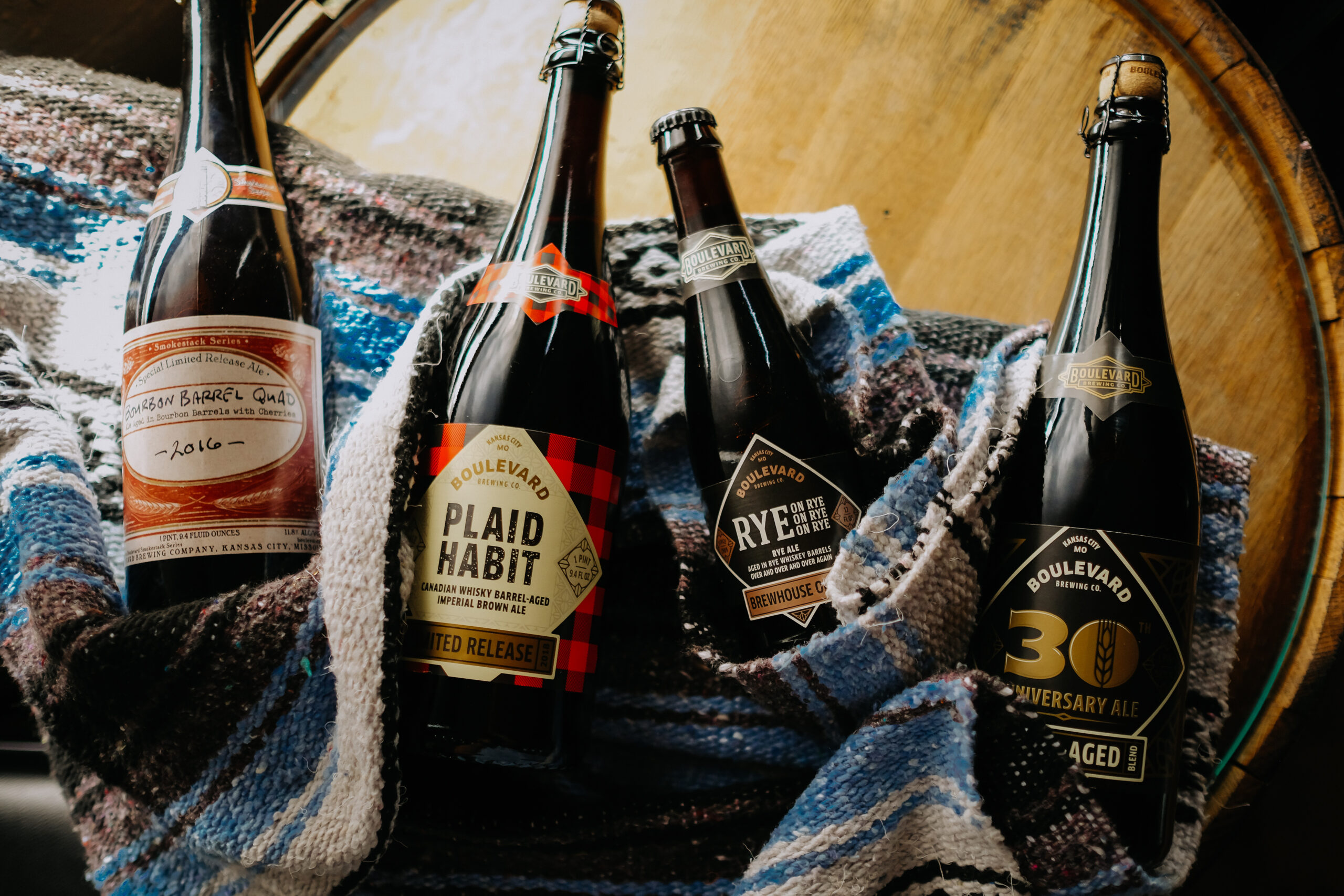 A collection of Boulevard beer bottles wrapped in colorful, woven blankets. The labels are visible, showcasing different varieties, including Victory Barrel Quad, Plaid Habit, Rye on Rye, and 30th Anniversary Ale. The background features a wooden barrel.