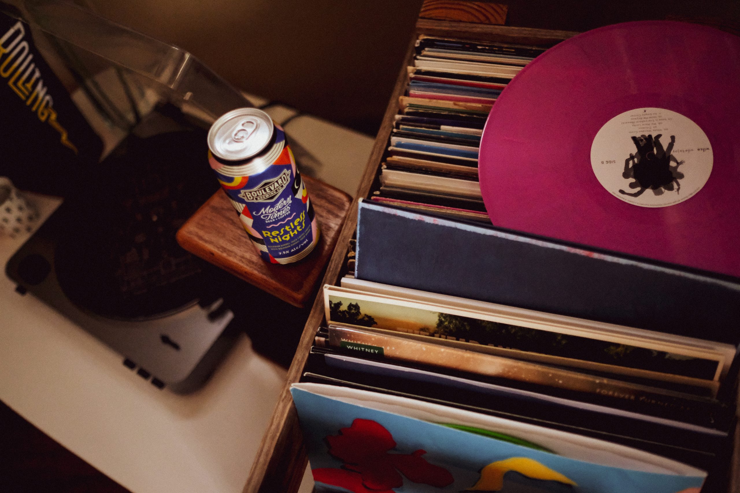 A vibrant magenta vinyl record on top of a stack, with a can of craft beer beside it on a wooden shelf. A vinyl player is seen partially in the background, and various album covers are visible in the stack.