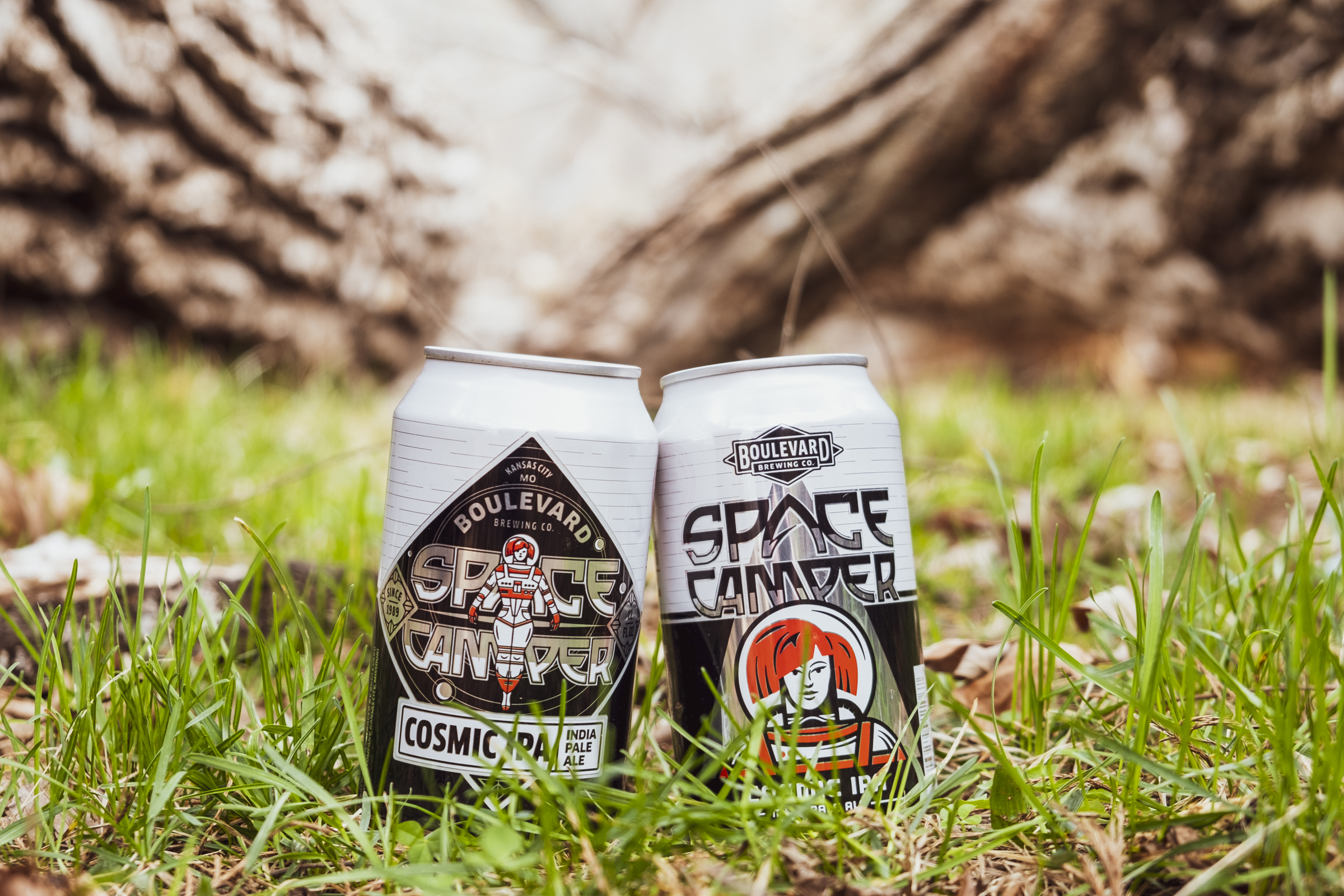 Two cans of Boulevard's Space Camper Cosmic IPA are placed on grass with a tree trunk in the background. The cans feature colorful, space-themed artwork, including an image of an astronaut on one can and a stylized cosmic design on the other.