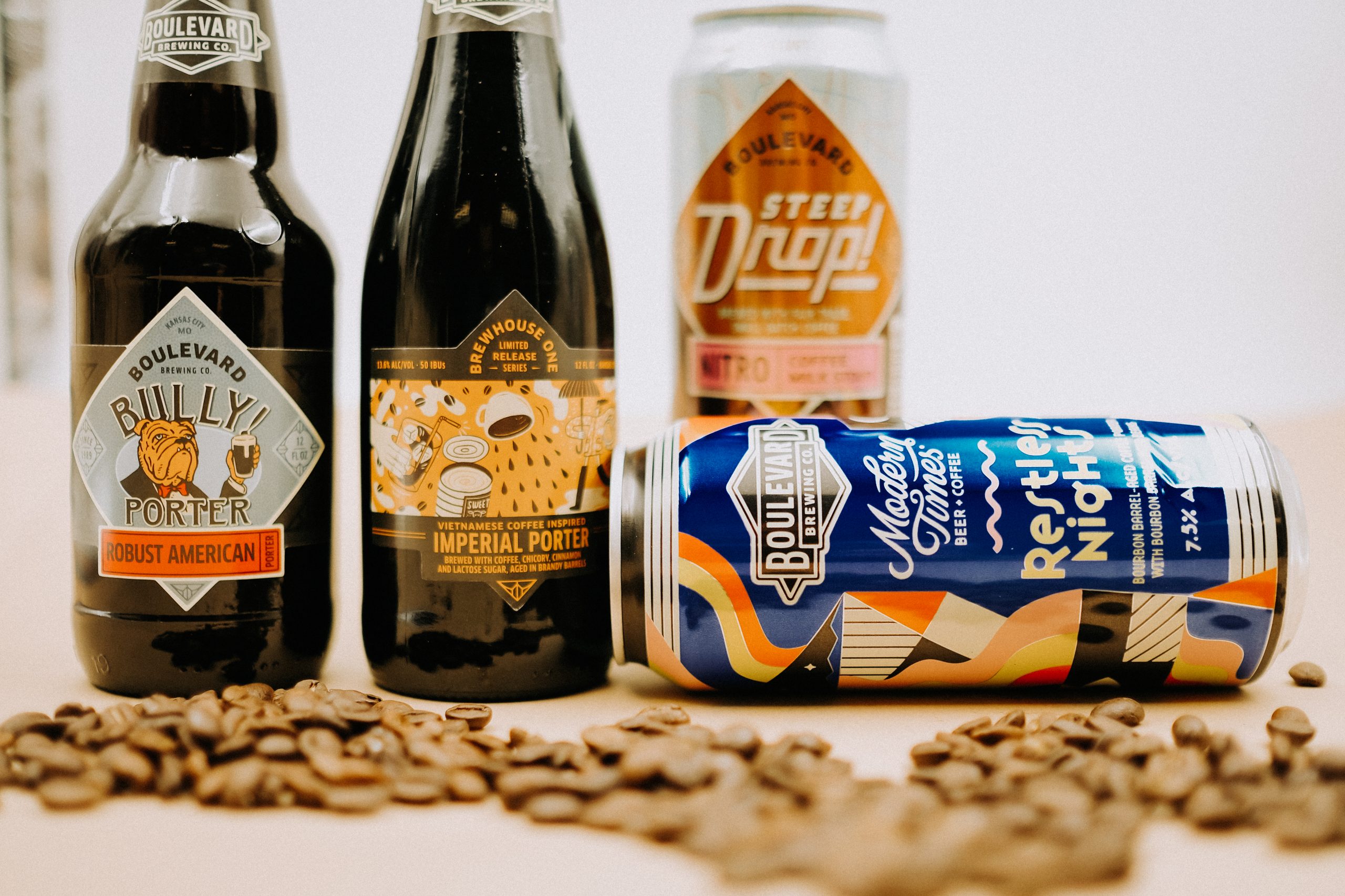 A variety of beer bottles and cans, including Billy's Porter and Imperial Porter, are displayed on a table surrounded by scattered coffee beans. The labels feature creative designs and branding elements.