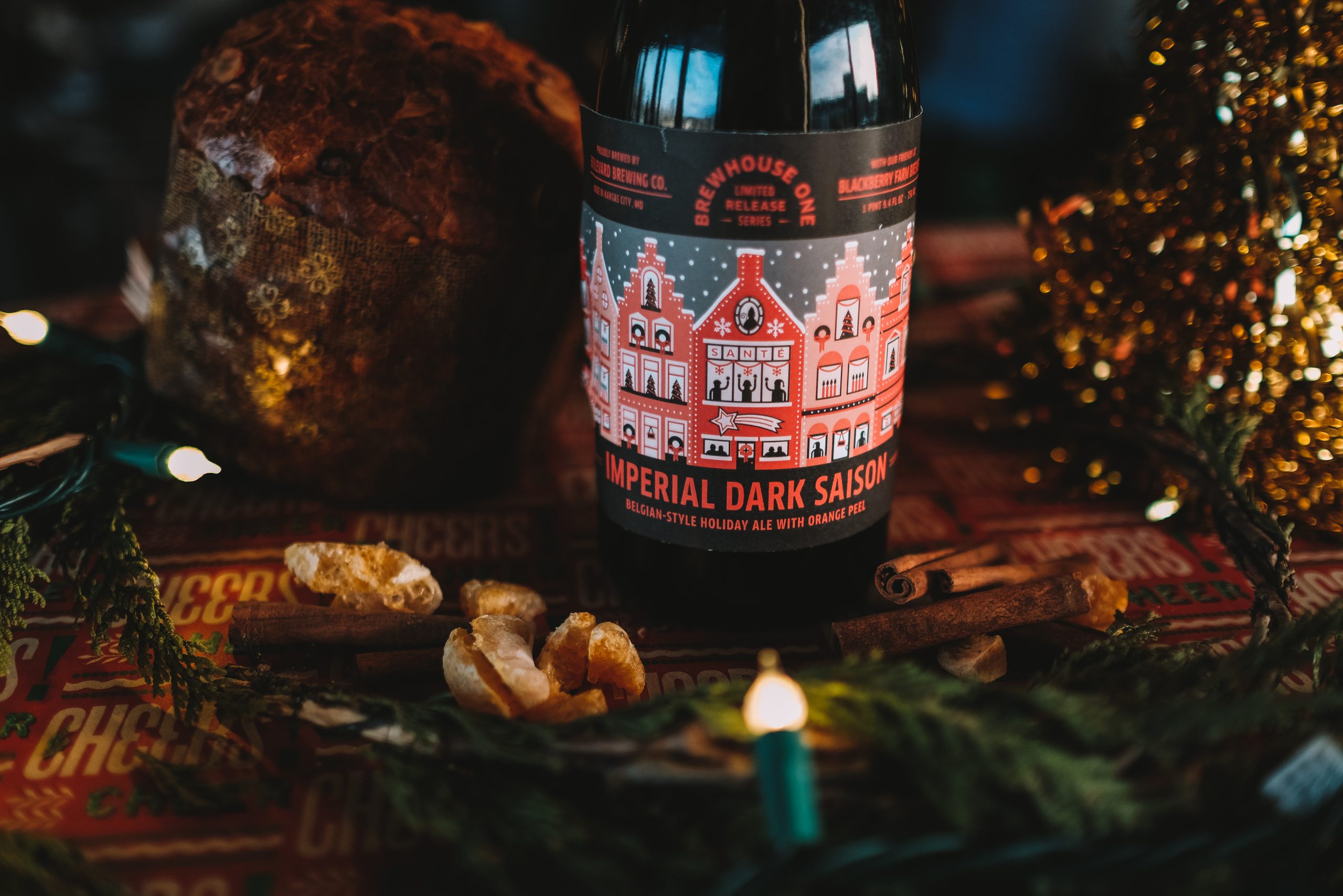 A festive holiday scene featuring a bottle of "Imperial Dark Saison" beer, surrounded by decorative greenery, holiday lights, cinnamon sticks, and a panettone. The beer label features a stylized illustration of traditional houses.