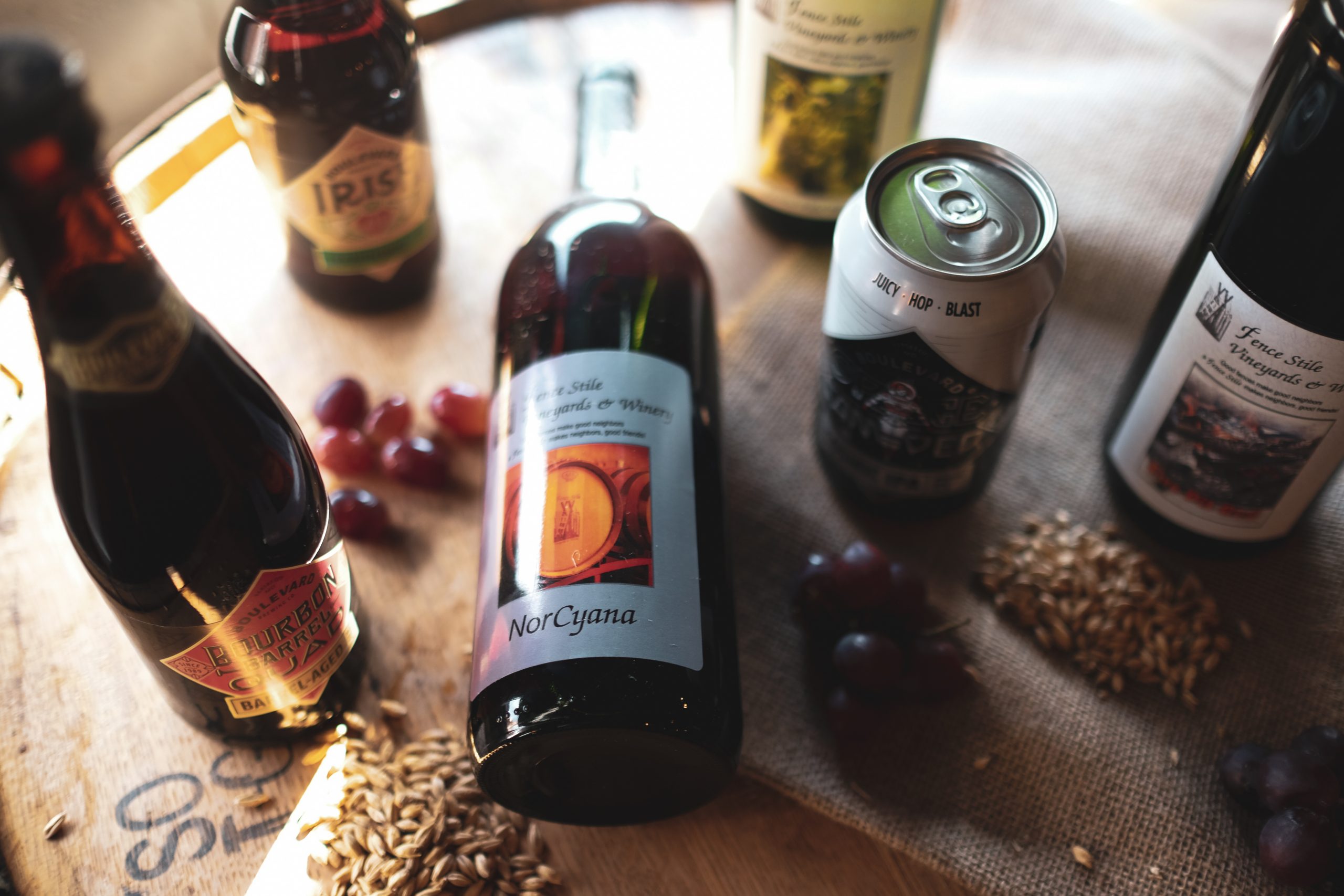 A variety of alcoholic beverages, including bottles and a can, are displayed on a wooden surface with scattered grapes and grains. One bottle is prominently placed in the center, labeled "NorCyana." The setting evokes a rustic and artisanal atmosphere.