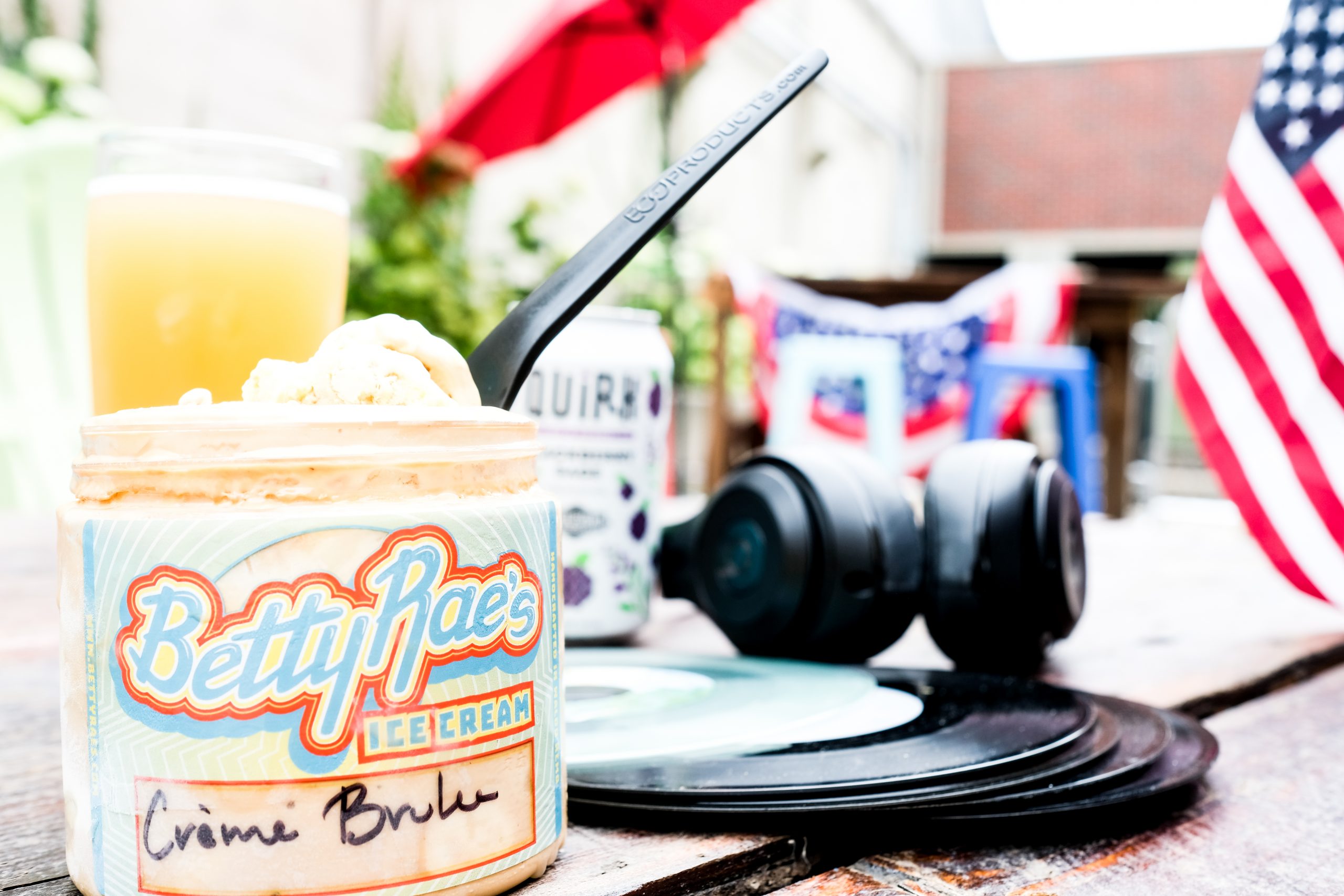 A Betty Rae's Ice Cream container sits on a wooden table with a spoon inserted in the ice cream. Nearby are a smartphone, a can of QUILT, a set of headphones, a stack of vinyl records, and an American flag. The setting appears to be an outdoor patio.