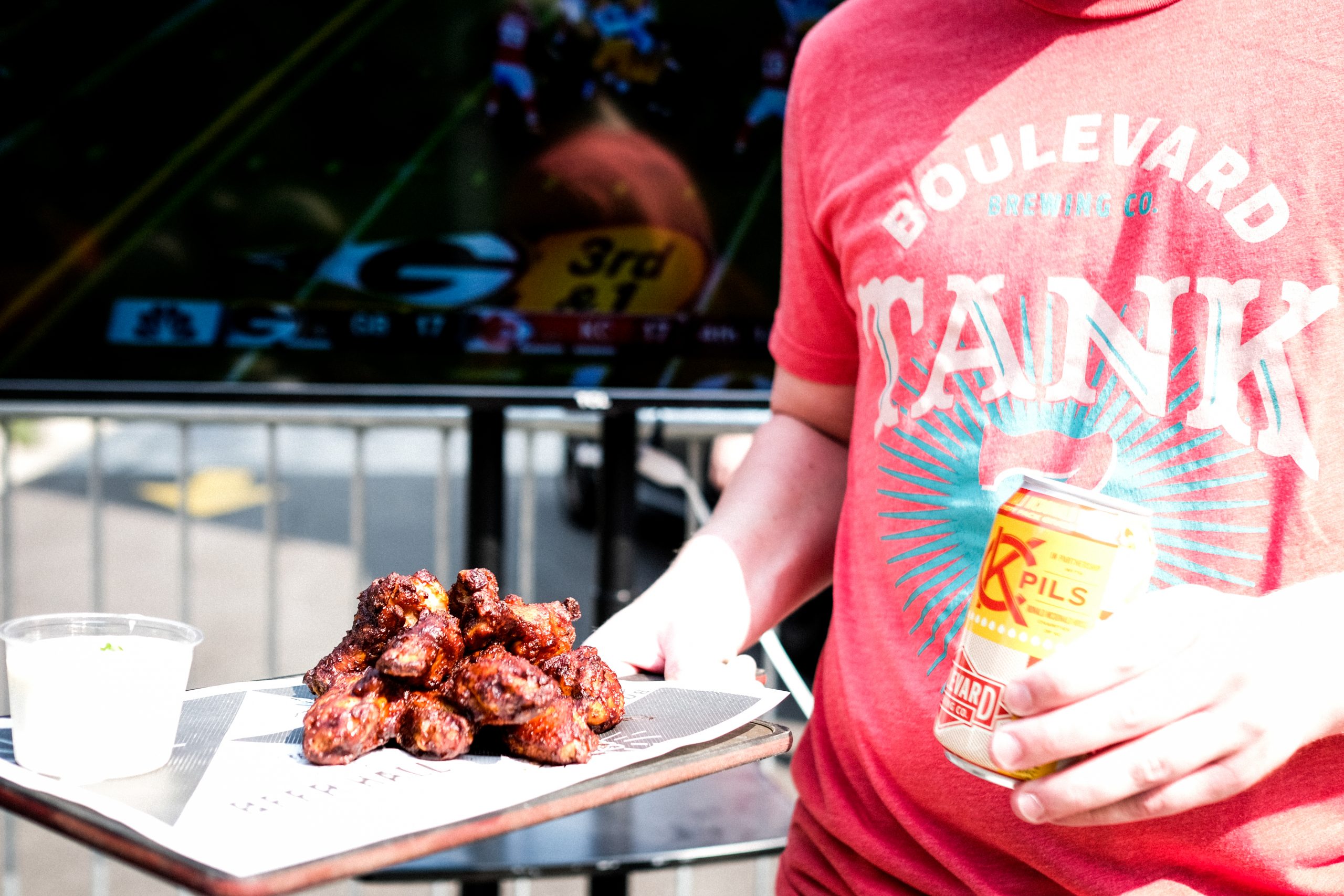 A person wearing a red "Boulevard Tank" shirt holds a tray of saucy chicken wings and a can of beer in front of a TV screen showing a football game. The bright sunlight emphasizes the food and drink.