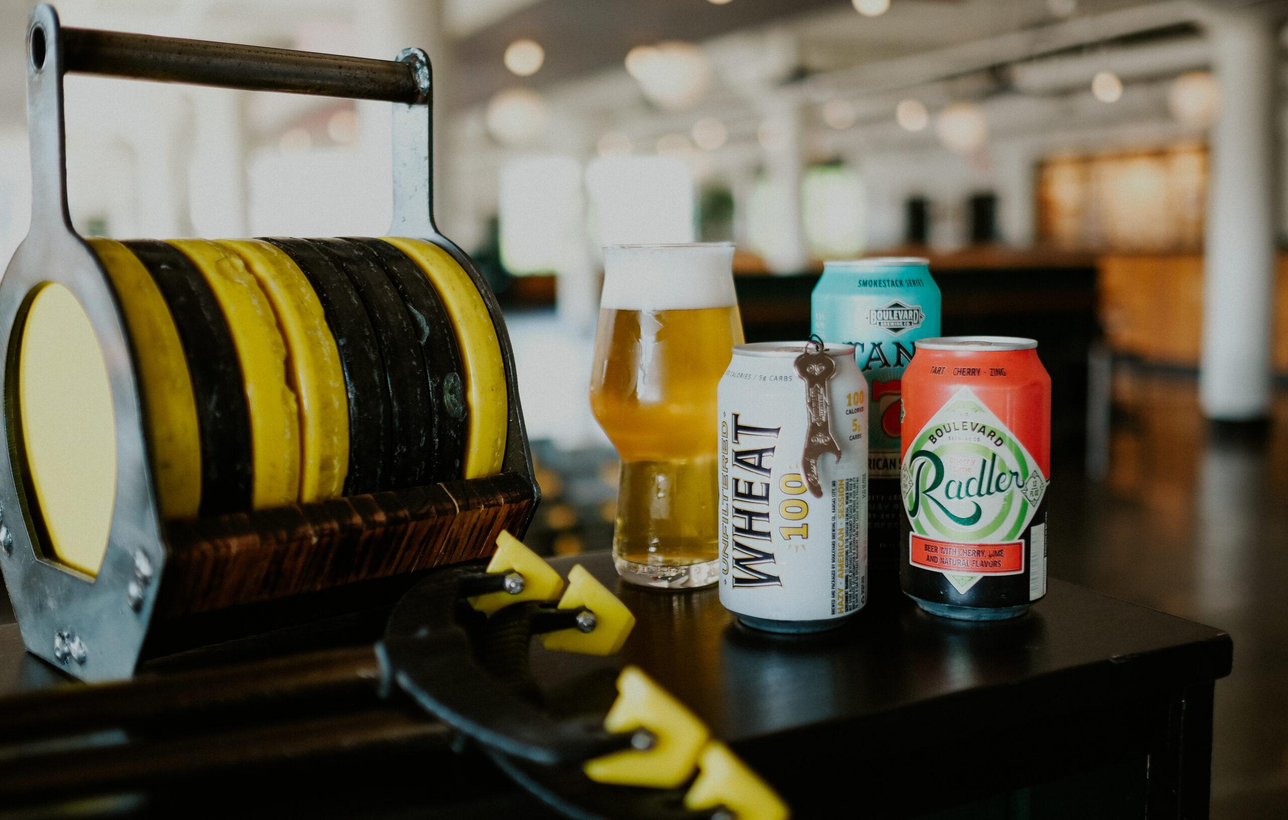 A table with a curling stone, a beer glass filled with beer, and three different beer cans. The cans are labeled as "Wheat 100," "Radler," and an ambiguous beer with turquoise and white design. The setting appears to be a casual indoor environment.