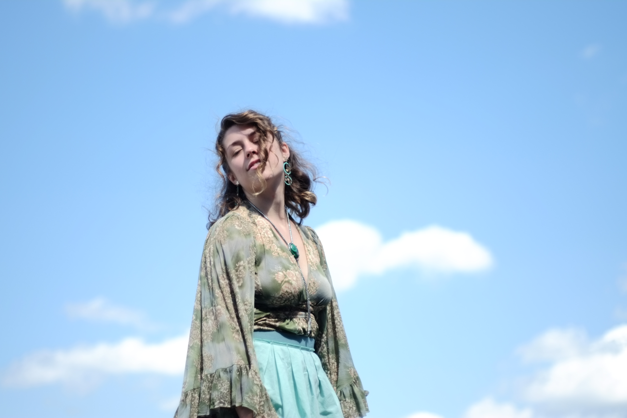 A woman with curly hair stands against a blue sky with scattered clouds. She is wearing a green floral blouse and a light blue skirt, with large earrings and a pendant necklace, and appears to be in a relaxed, serene pose.