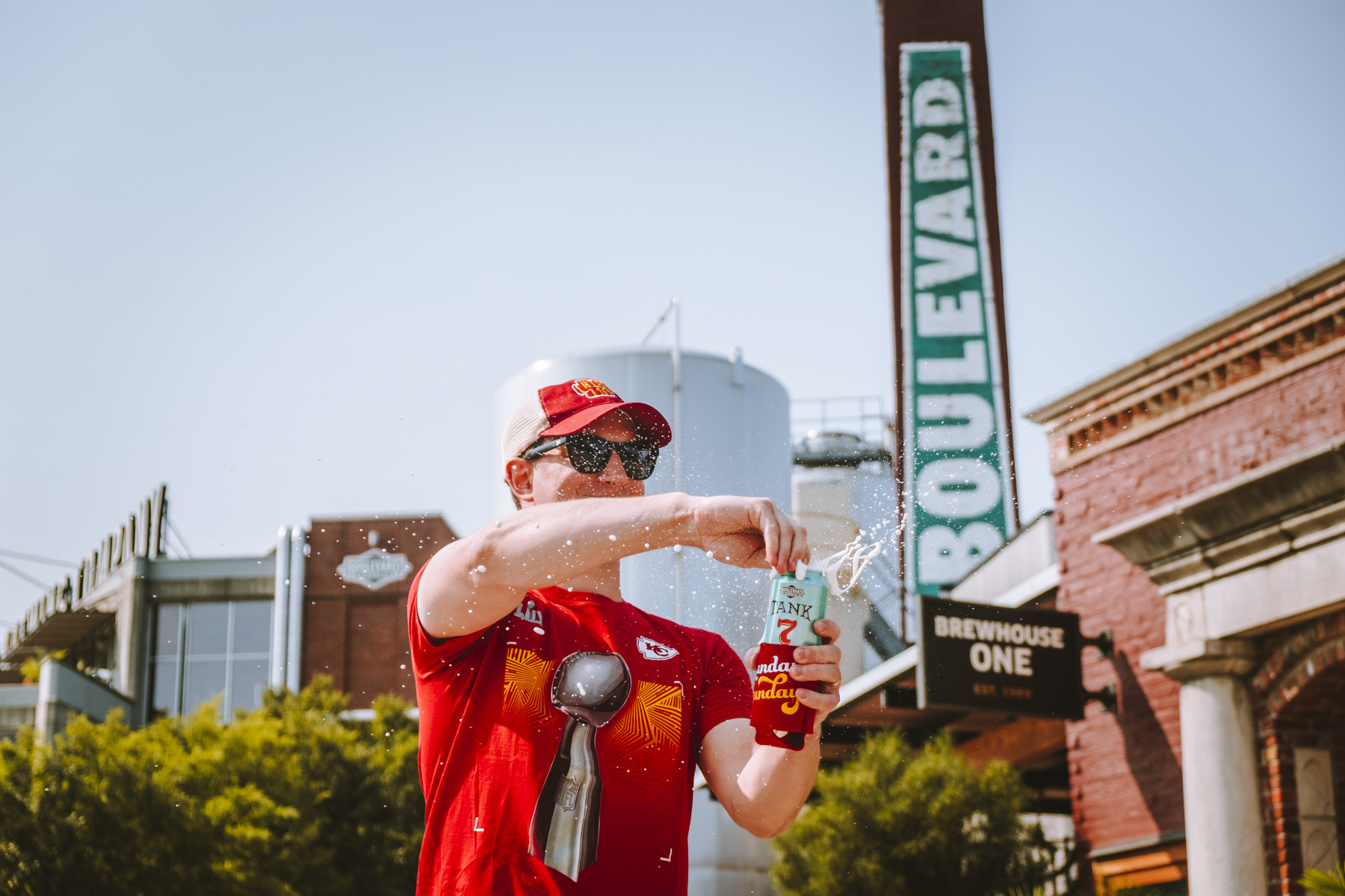 A person wearing a red shirt, cap, and sunglasses sprays a can of beer while standing in front of a building with a sign that reads "BOULEVARD" and another sign saying "BREWHOUSE ONE." There is a tank and greenery in the background.