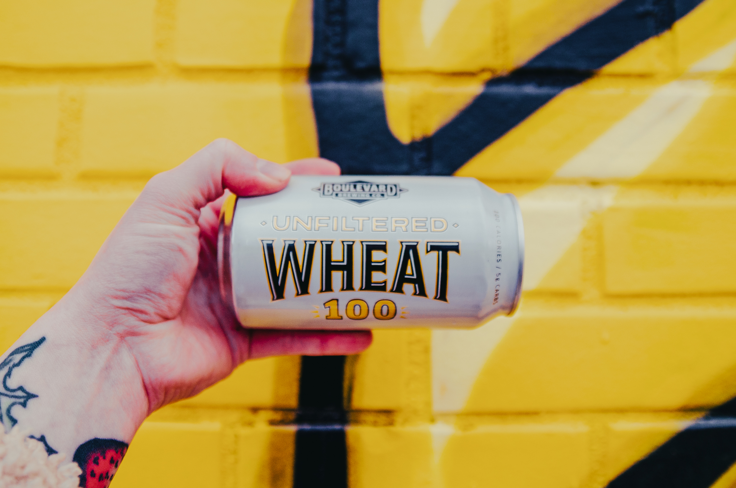 A hand with a tattoo on the wrist is holding a can of Boulevard Unfiltered Wheat 100 beer. The background is a yellow painted brick wall with black and yellow abstract shapes.