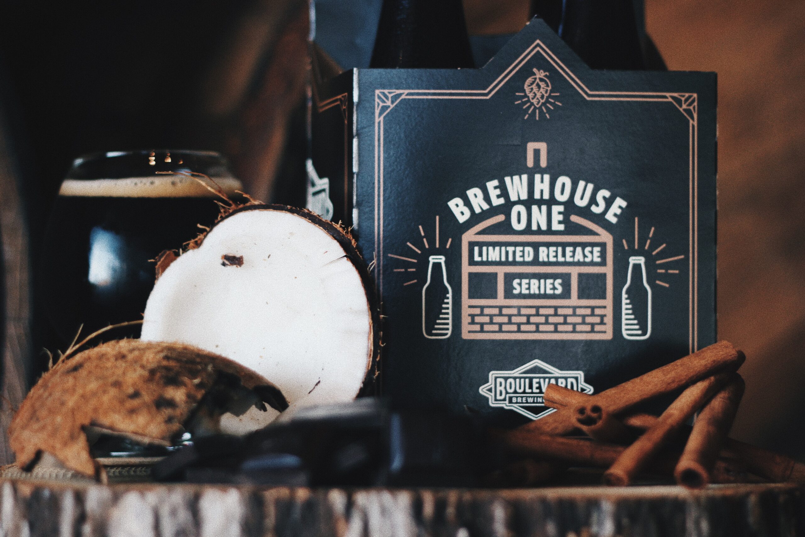 A close-up of a Brewhouse One limited release beer package with a dark beer glass beside it. Coconut halves and cinnamon sticks are artistically arranged in the foreground.