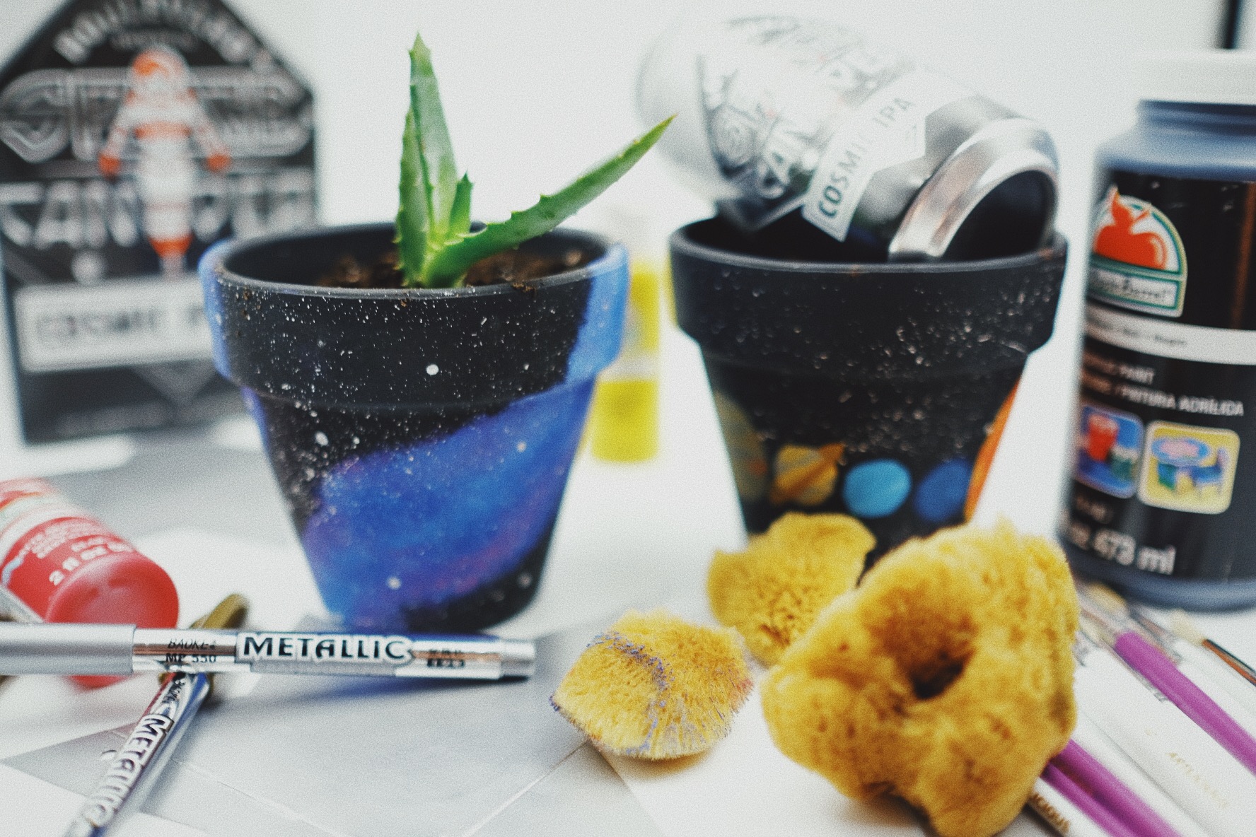 A craft scene with black painted pots featuring galaxy and planet designs. One pot has a small plant, the other holds a beverage can. Art supplies including paints, brushes, and sponges are scattered around.