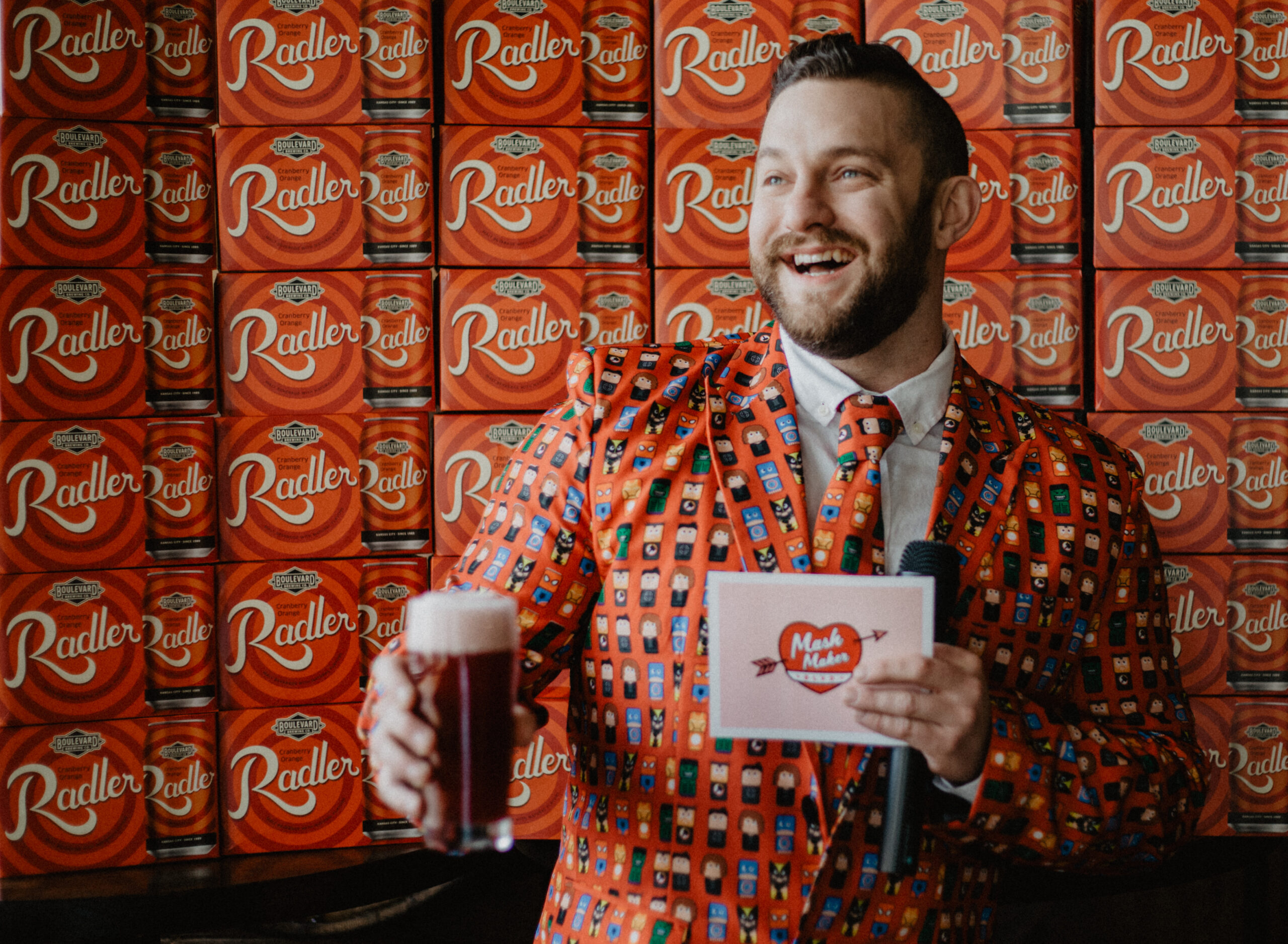 A man in a colorful, patterned suit holds a microphone and a drink, smiling joyfully. He's standing in front of a wall stacked with red Radler beer boxes.