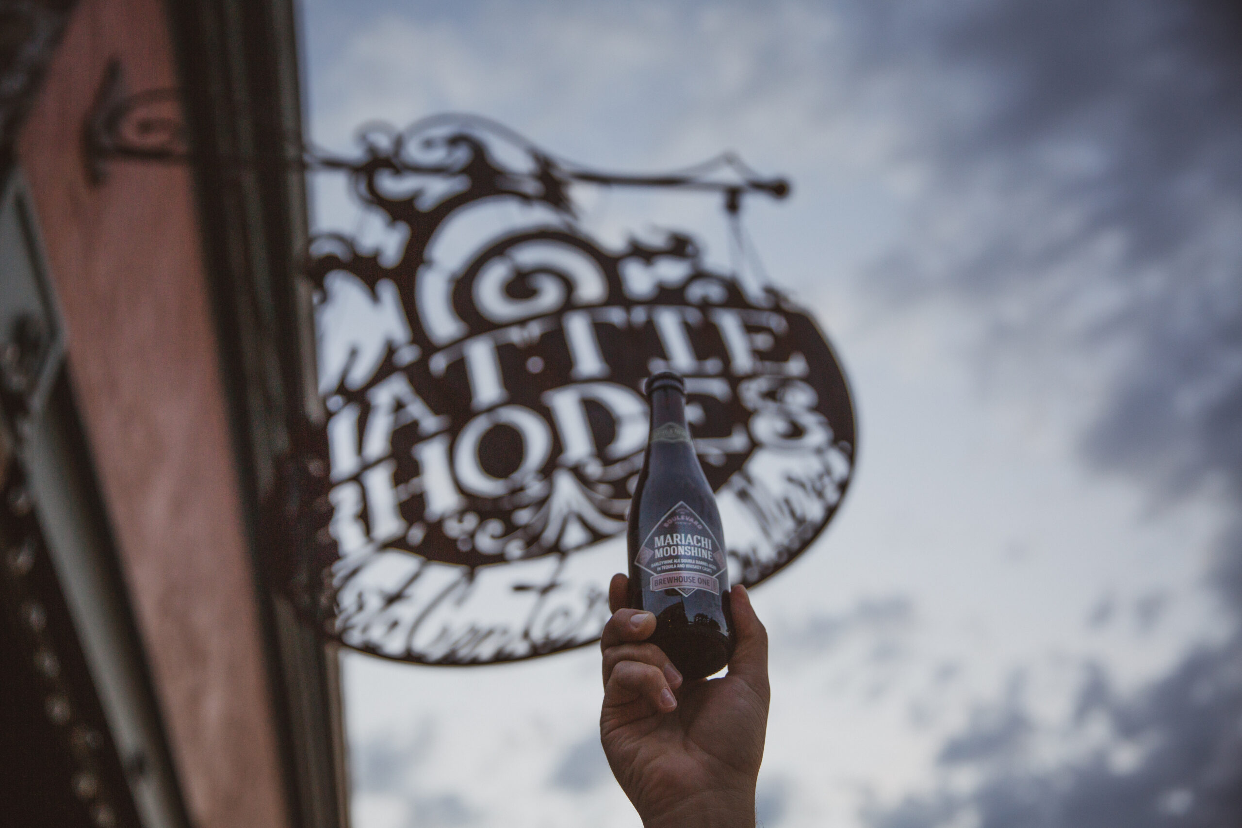 A hand holds up a dark bottle of beer against a cloudy sky. In the background, there's a hanging sign that appears to read "Mattie Rhodes.