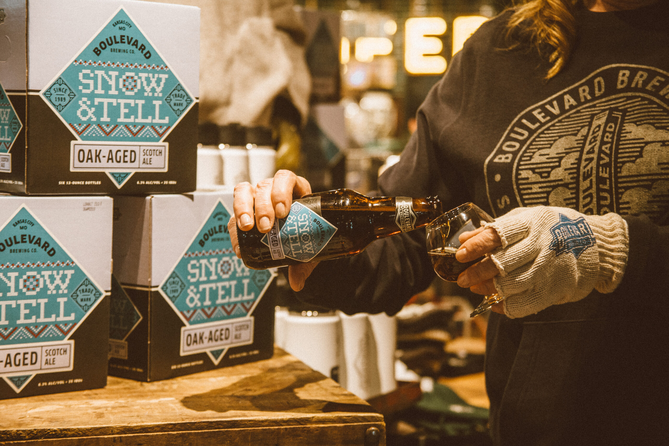 A person wearing a sweater and gloves pours a bottle of Boulevard Brewing Co.'s Snow & Tell Oak-Aged Scotch Ale into a glass. Several boxes of the ale are stacked nearby. The setting appears to be a cozy, warmly lit indoor space.
