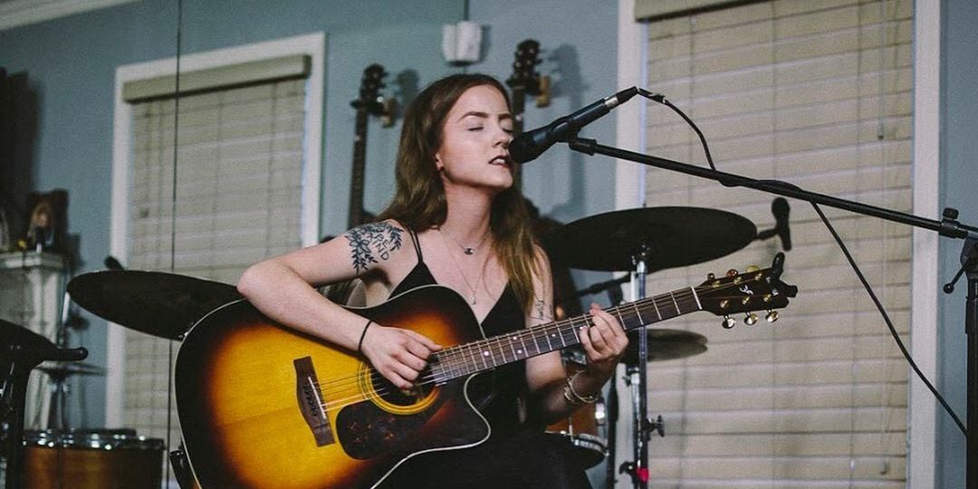 A woman with long brown hair and a tattoo on her arm plays an acoustic guitar and sings into a microphone. She is seated in front of a drum set, and the setting appears to be a cozy indoor space with blinds on the windows.