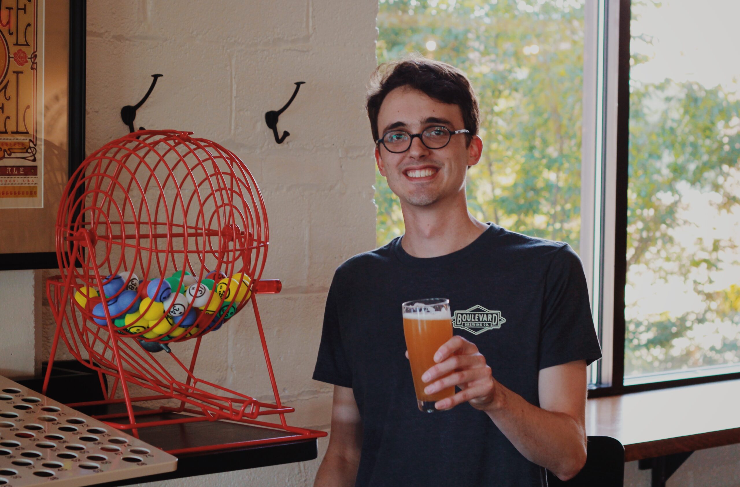 A person wearing glasses holds a glass of beer and stands next to a red cage filled with colorful balls. They are smiling, with a window showing a green tree in the background.
