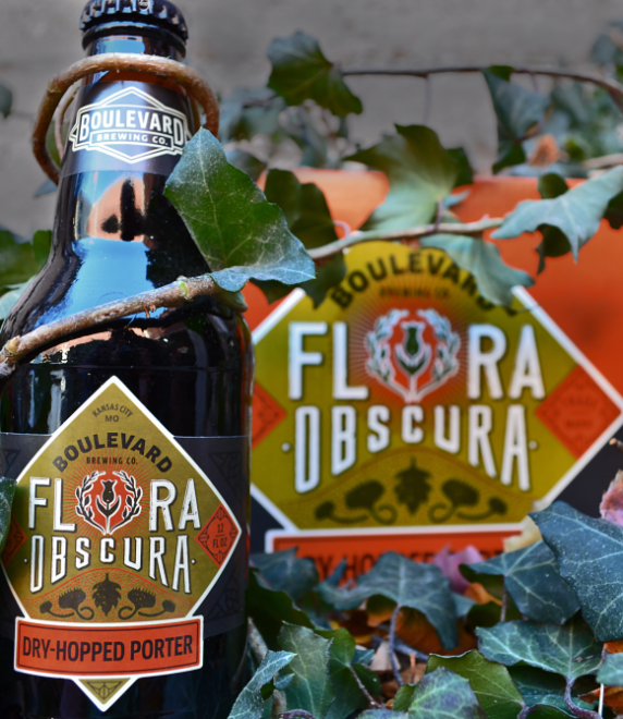 A bottle of Boulevard Brewing Co.'s Flora Obscura Dry-Hopped Porter is surrounded by autumn leaves. The bottle labels feature a decorative design with a hexagonal logo. The colors are rich and earthy.