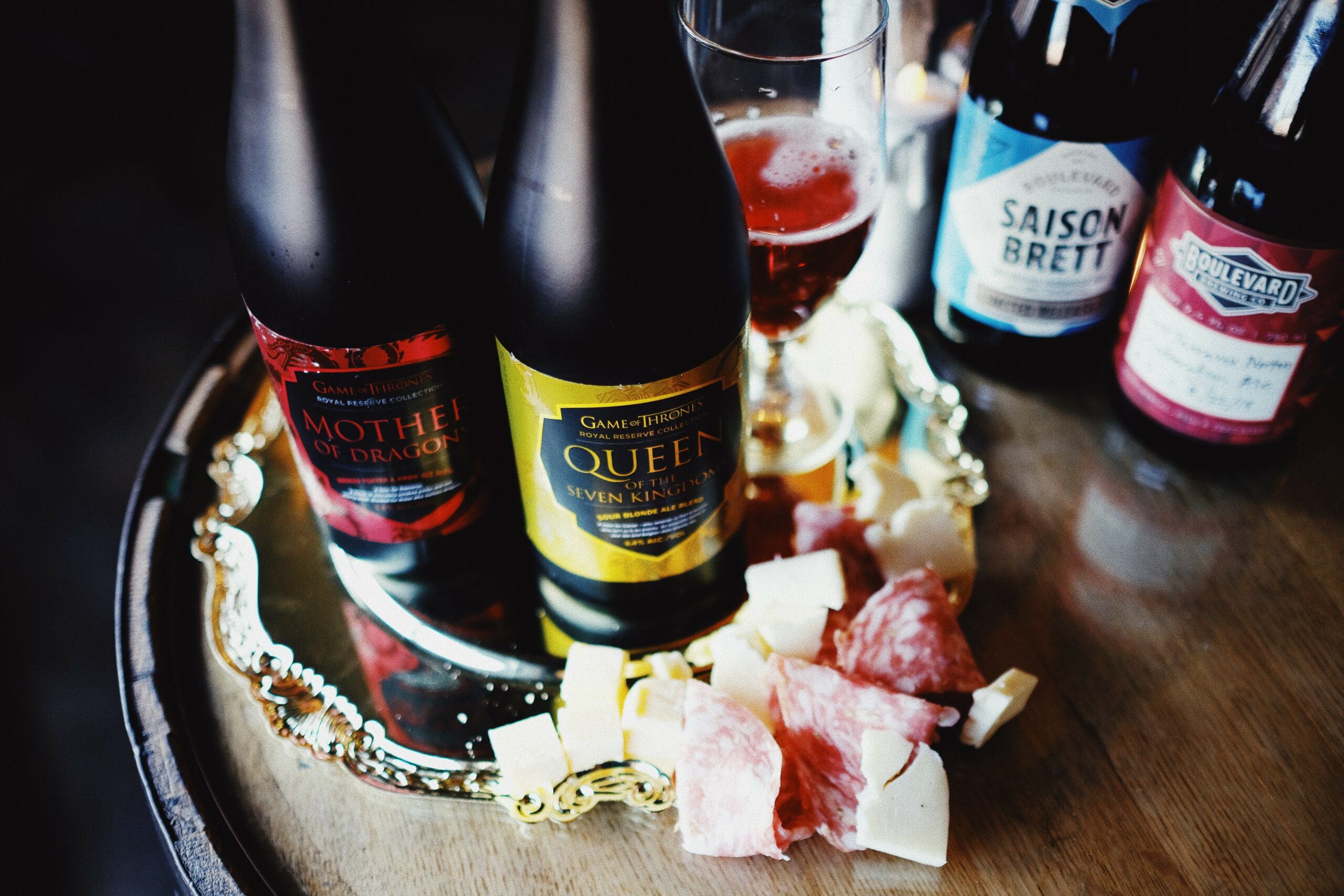 A glass of red beer, two bottles of themed craft beer, and other assorted beer bottles are displayed on a reflective tray. Nearby are small slices of cheese and cured meat on a wooden surface.