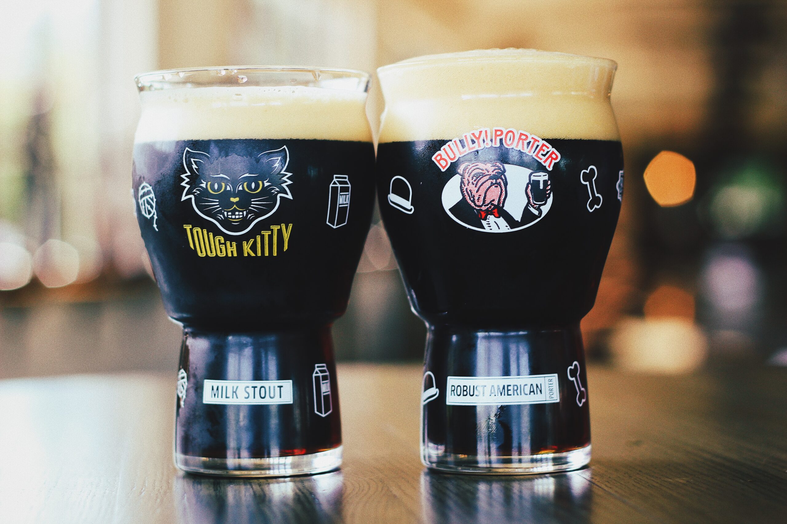 Two glasses of dark beer on a table, one labeled "Tough Kitty Milk Stout" with a cat illustration, the other "Bulky Porter Robust American" featuring a weightlifting character. The background is blurred with warm lighting.