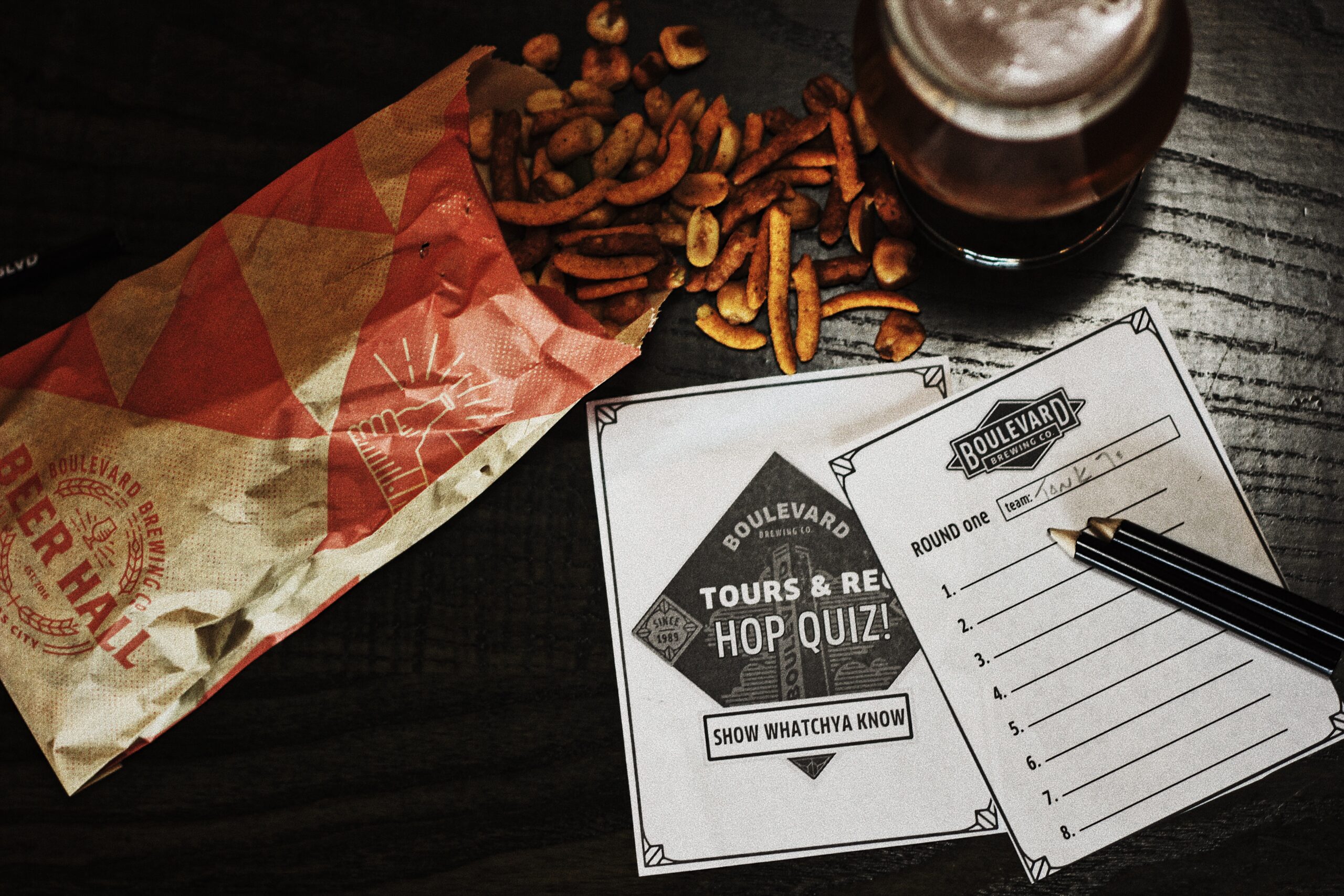 A tabletop scene with a crumpled snack bag, scattered nuts, a glass of amber beer, and a trivia quiz sheet with the title "Tours & Rec Hop Quiz!" labeled "Boulevard." A pen rests on the sheet beside a numbered answer section.