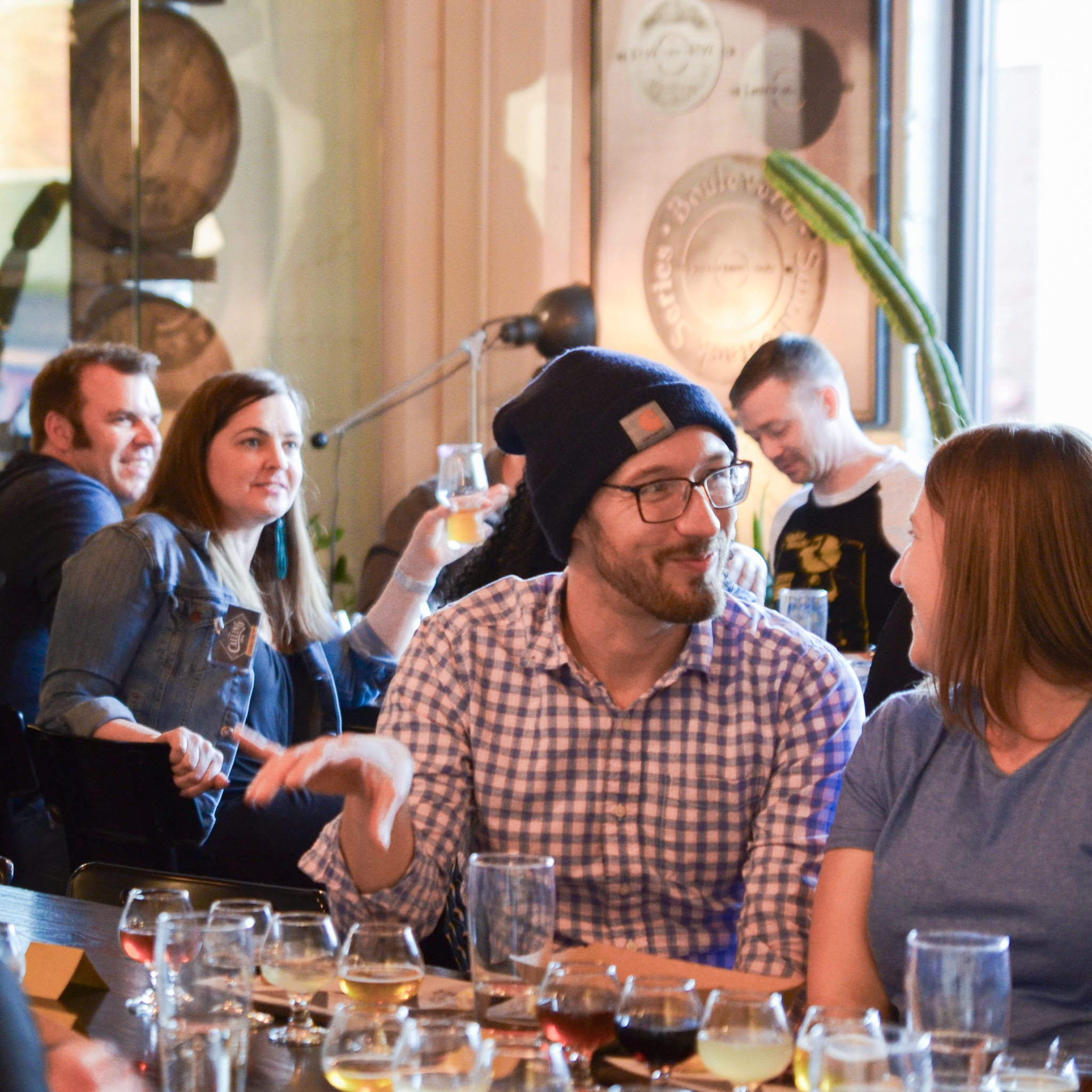 A group of people sit at a bar, engaging in conversation and enjoying drinks. A man in a checkered shirt and beanie gestures while talking to a woman in a blue shirt. There are several glasses on the table, and a large barrel is visible in the background.