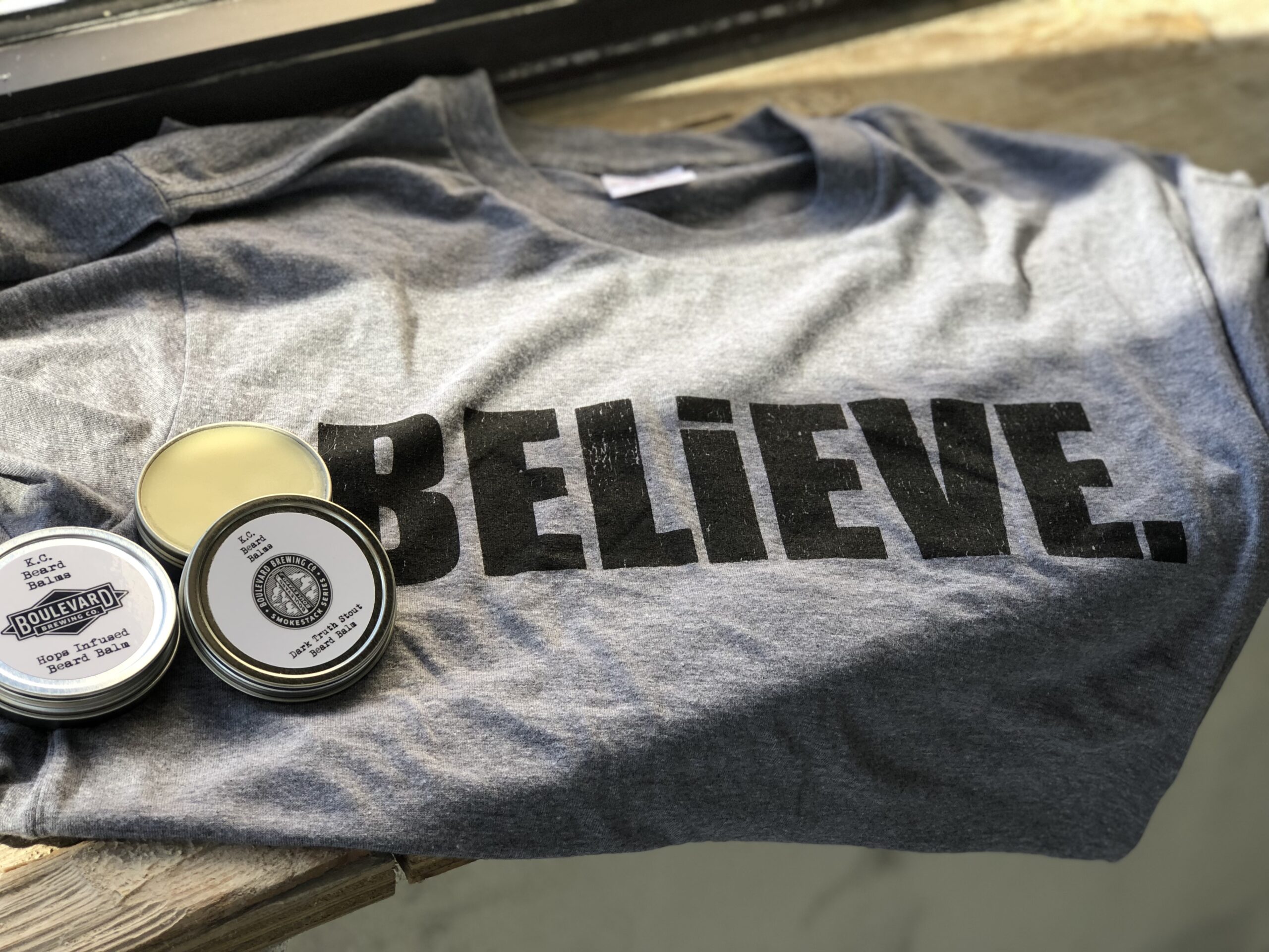 A gray T-shirt with the word "BELIEVE." printed in bold black letters is laid out on a wooden surface. Next to it are two small tins, partially open, containing balm or cream. Light filters through a nearby window.