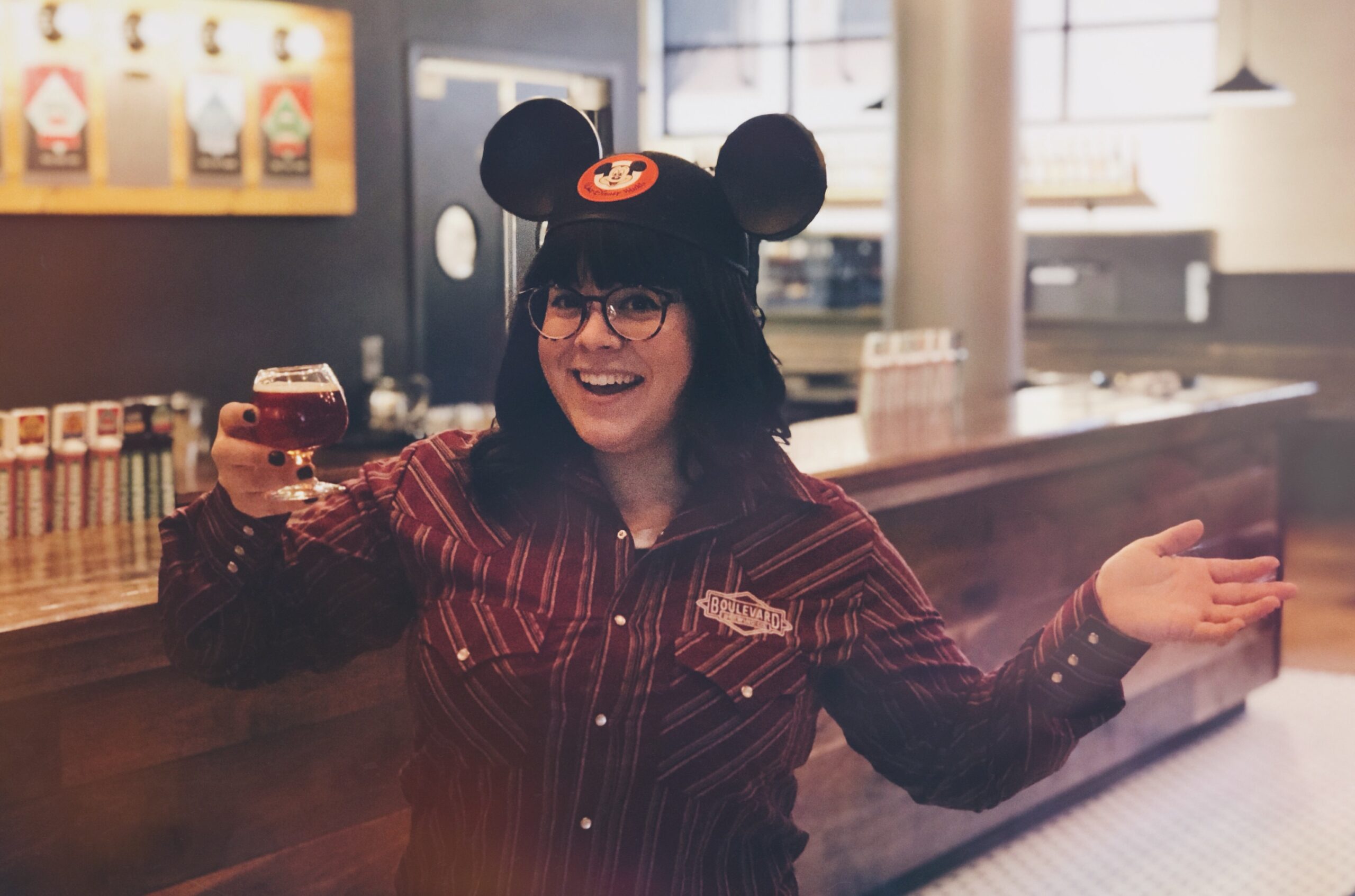 A person wearing Mickey Mouse ears smiles broadly while holding a glass of beer. They stand in a warmly lit room with a wooden counter and taps in the background.