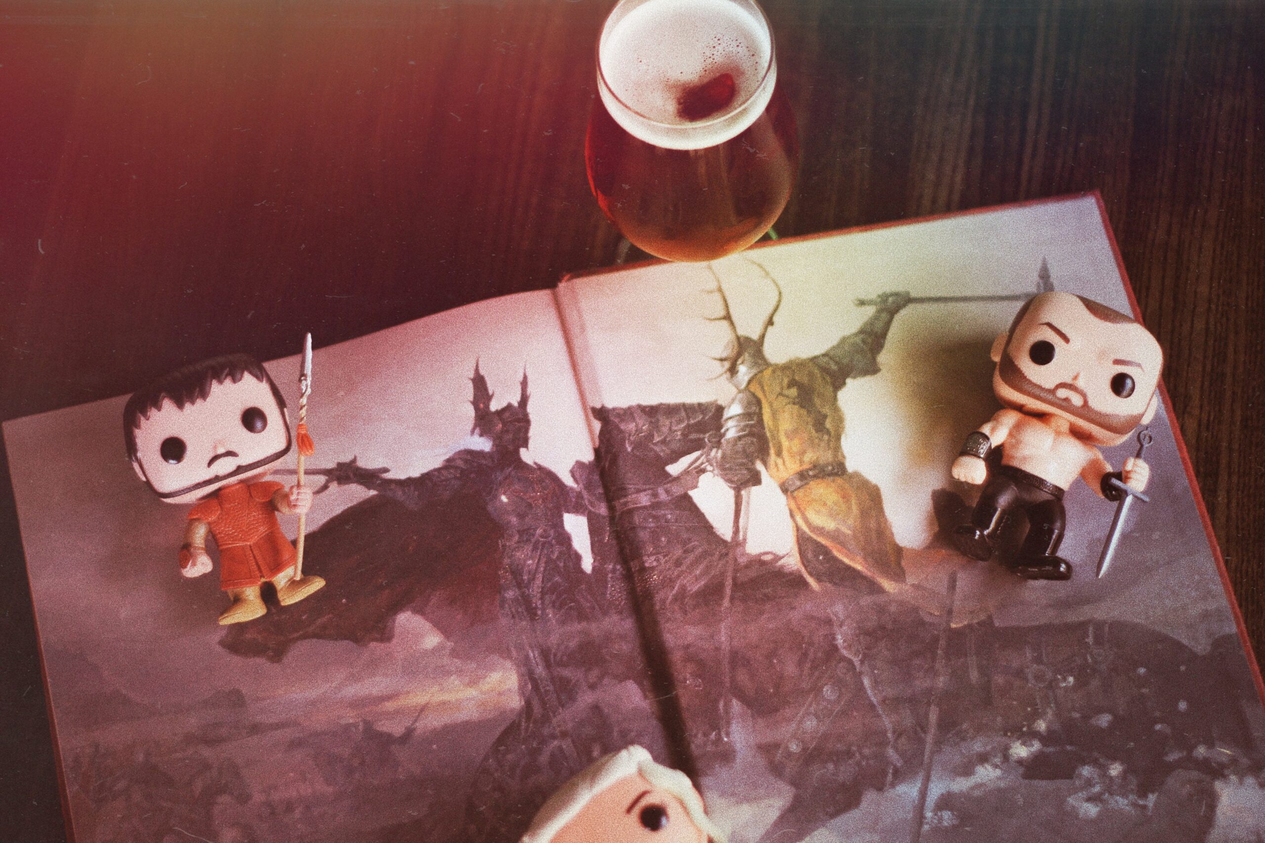 Two figurines with swords are placed on an open fantasy book depicting warriors. A glass of dark beer is positioned above the book.