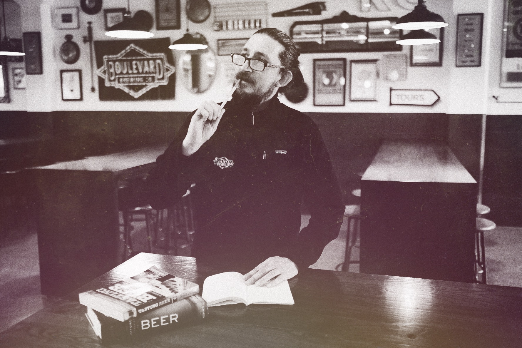 A man with glasses stands thoughtfully at a wooden table in a vintage-style brewery. He holds a pen and has an open book and two books titled "Beer" in front of him. The room has a nostalgic decor with black and white photography.