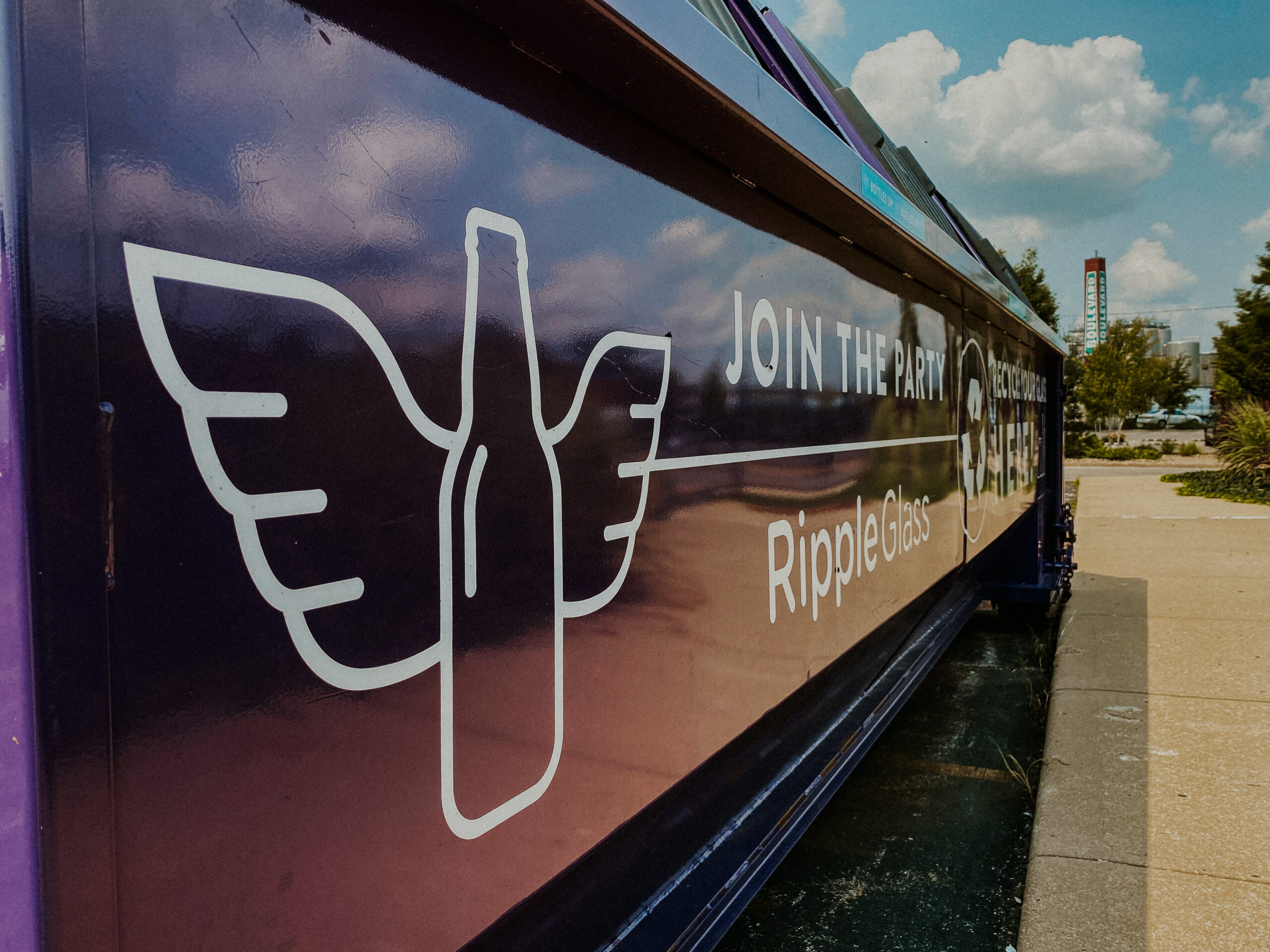 Close-up of a purple recycling container with a white graphic of a bottle with wings and the text "Join the Party" and "Ripple Glass." The container is outdoors on a sunny day, with trees and a clear sky in the background.