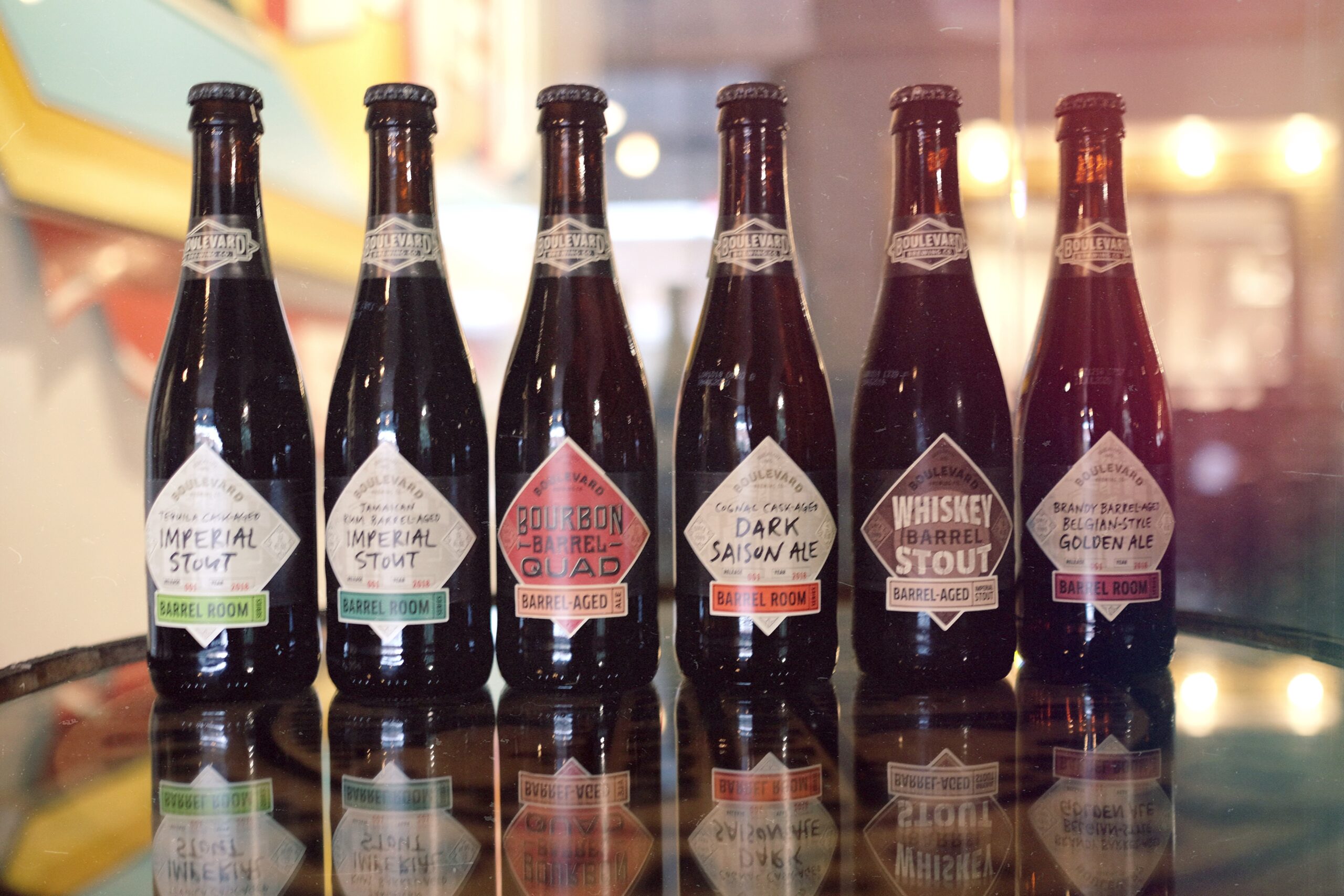 Six dark beer bottles are displayed in a row on a reflective surface. Each bottle features a unique label with different types of stout and aged beer, including Imperial Stout, Bourbon Barrel Quad, and Whiskey Barrel Stout.