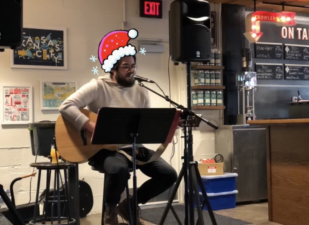 A person sits on a stool playing an acoustic guitar and singing into a microphone. They wear a white sweater and have a cartoon Santa hat added above their head. The background features beverage displays and a chalkboard menu.