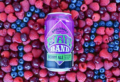 A can of Boulevard Jam Band Berry Ale rests on a bed of mixed berries, including raspberries, blueberries, and cherries. The can features a vibrant purple design with "JAM BAND" prominently displayed.