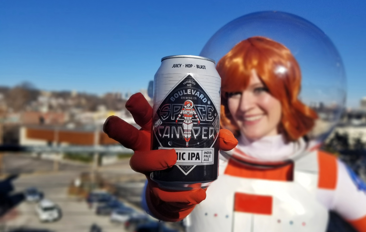 A smiling person in a space-themed costume holds a can of "Space Camper Cosmic IPA" by Boulevard Brewing. The background shows a blurred cityscape under a clear blue sky.