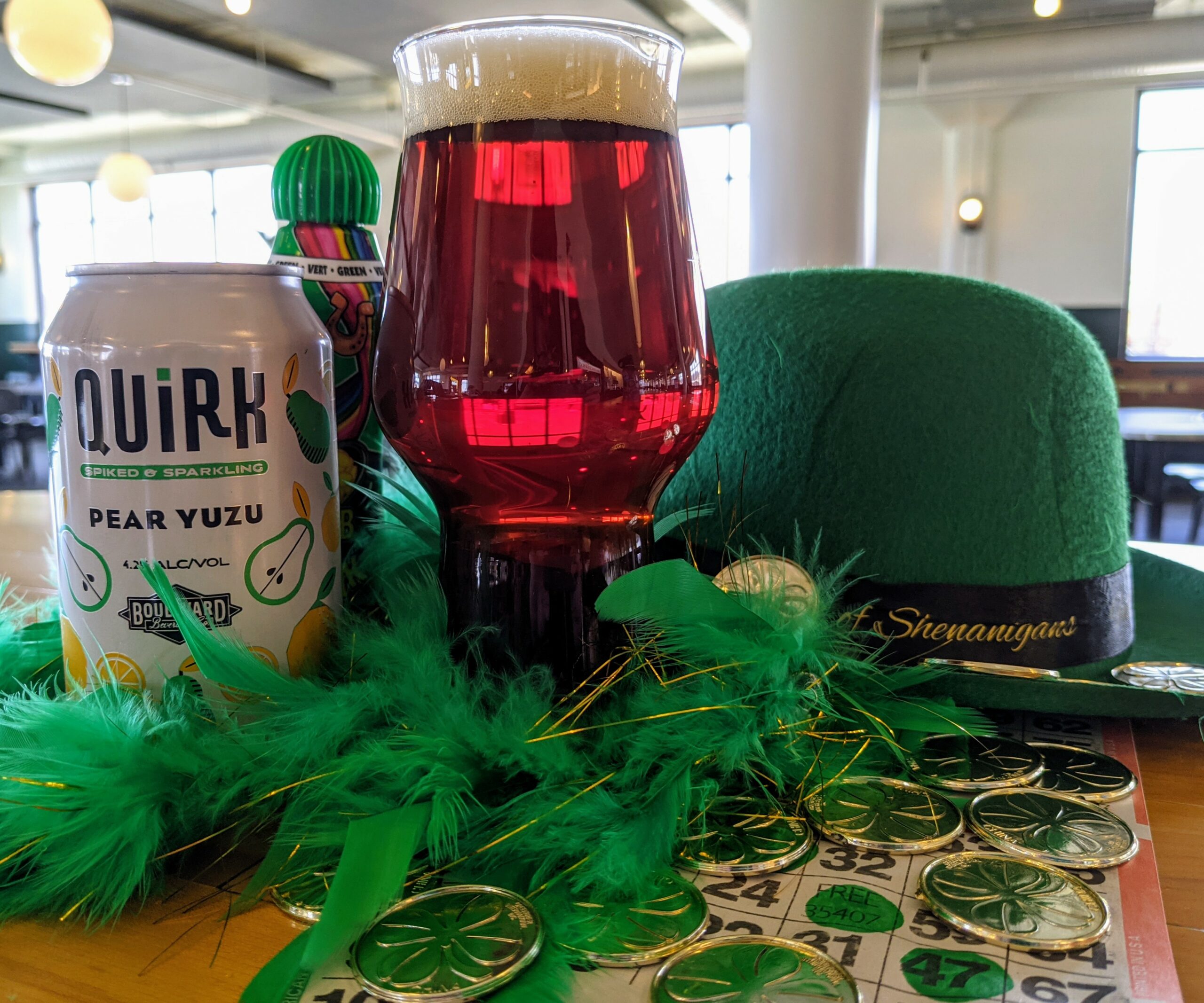 A table with St. Patrick's Day-themed items including a green hat labeled "Shenanigans", green feathers, gold coins, and a Quirk Pear Yuzu hard seltzer can. A glass of dark red beverage is placed at the center. Bingo cards and game pieces surround the setup.