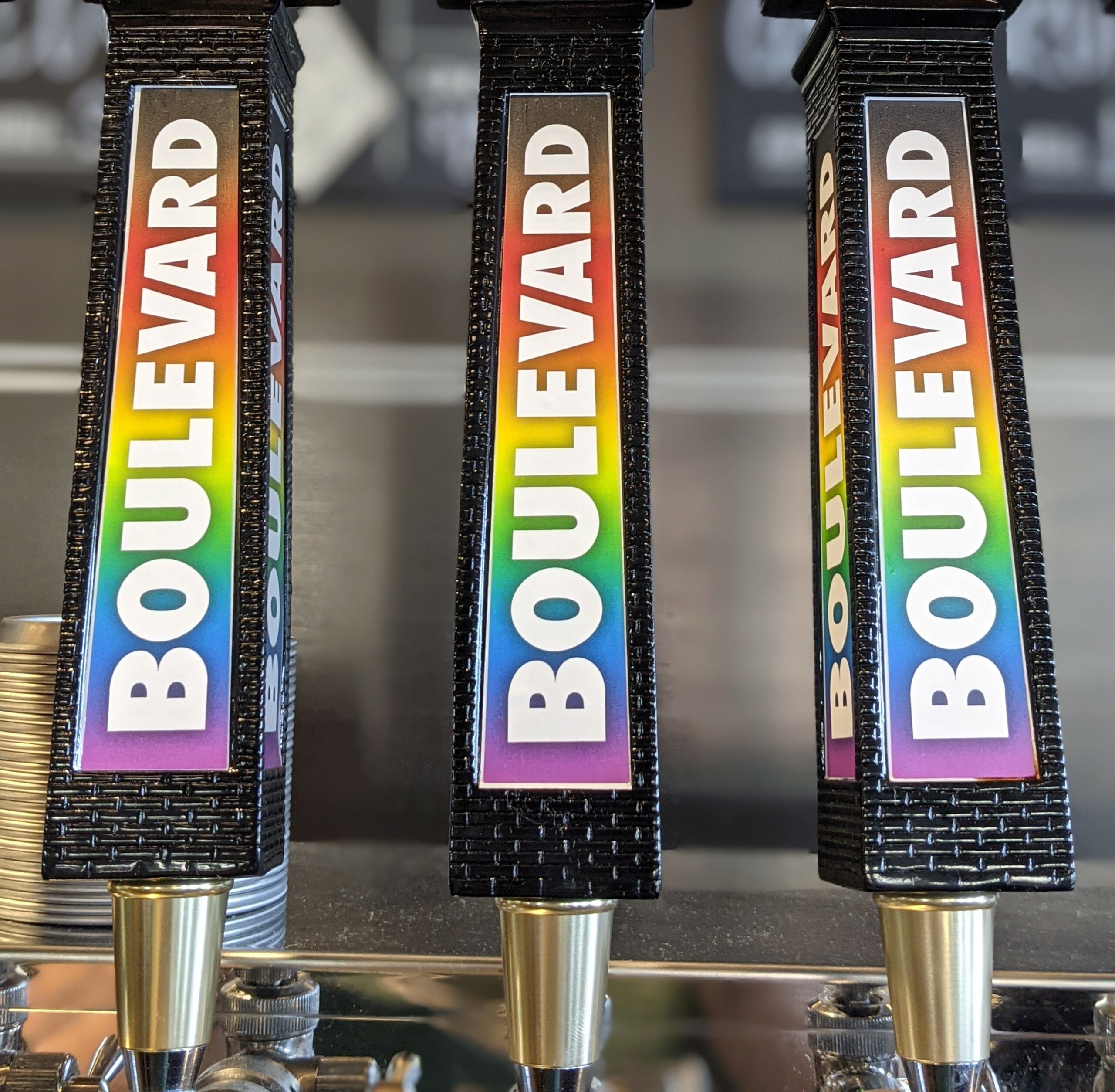 Close-up of three black beer tap handles labeled "Boulevard" with rainbow-colored text, showcasing the taps in a bar setting. The background is slightly blurred, highlighting the colorful branding of the beer handles.