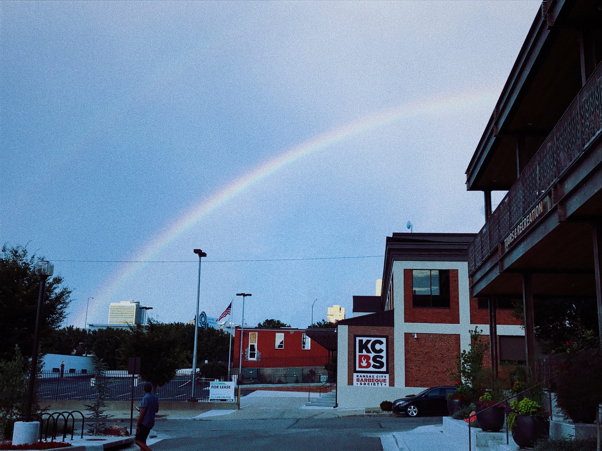 A faint rainbow stretches across a cloudy sky above a city street. A man walks on the sidewalk near a multi-story building and a smaller red brick structure with the letters "KCS" on the wall. Trees and lampposts line the street.