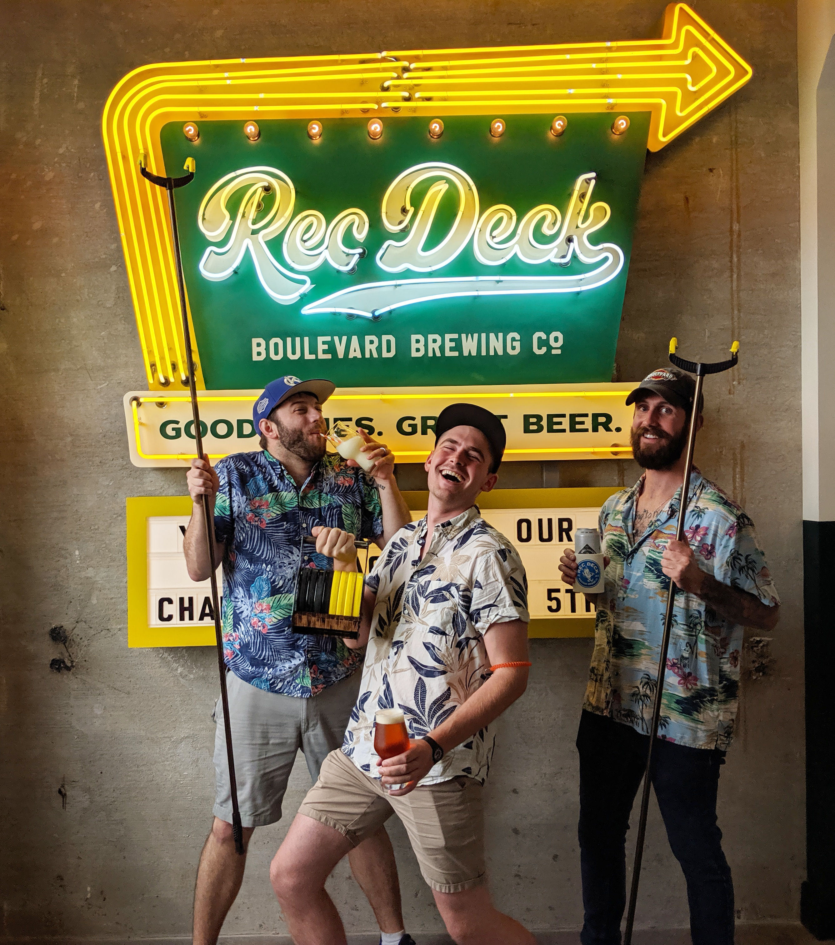 Three men in tropical shirts hold novelty golf clubs, posing energetically in front of a neon sign that reads "Rec Deck Boulevard Brewing Co: Good Times. Great Beer.