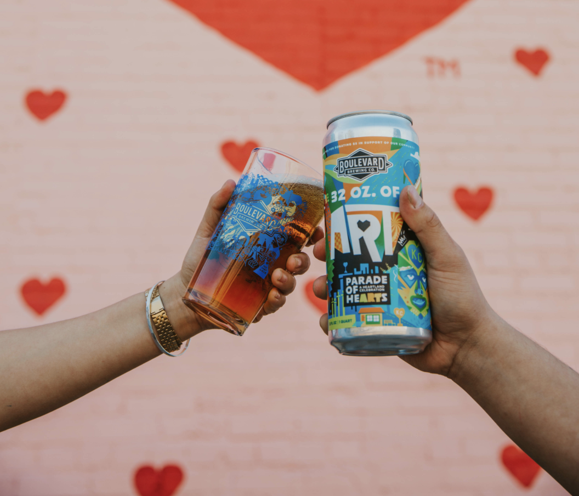 Two hands are clinking a pint glass and a can of beer in front of a pink brick wall with red hearts painted on it. The pint glass is filled and also displays a colorful design, matching the vibrant, patterned can. The background features a large red triangle.