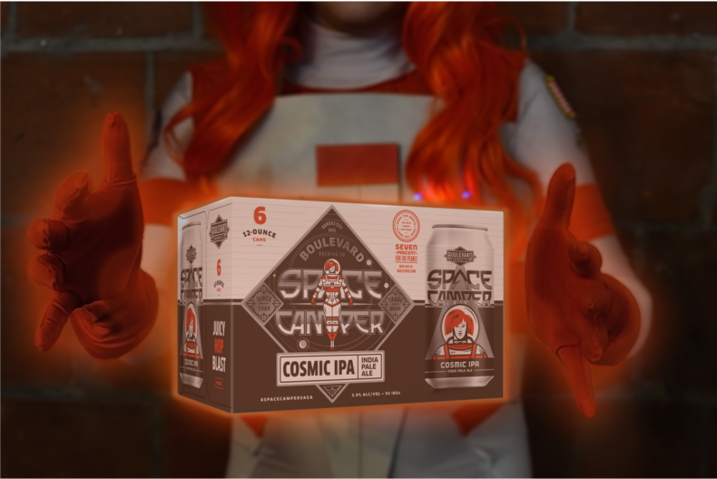 A person wearing an orange and white astronaut costume holds a six-pack of Boulevard Brewing Co.'s "Space Camper Cosmic IPA". The six-pack's label features a sci-fi themed design with an astronaut character. The background is a brick wall.