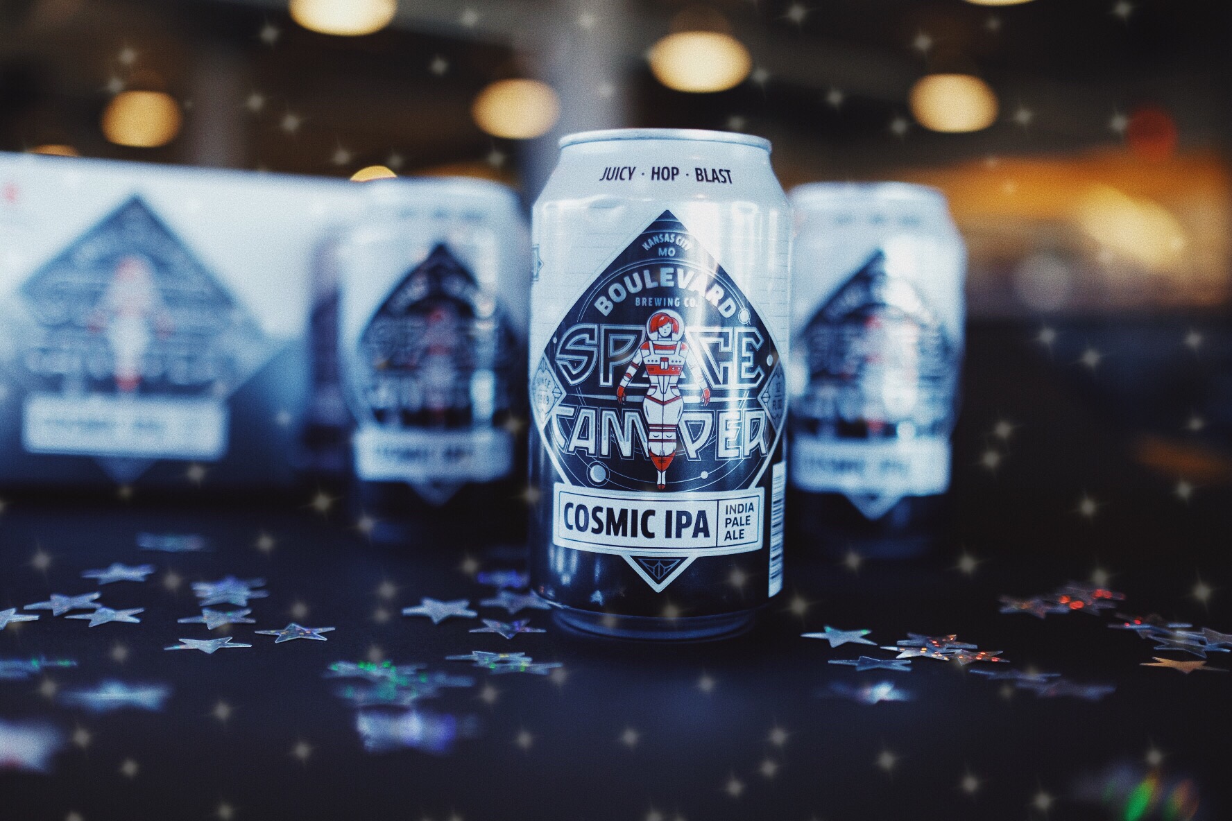 A can of Boulevard Space Camper Cosmic IPA is placed on a table with scattered star-shaped confetti. The can features bold, graphic artwork with space-themed design elements. Other cans and a box are blurred in the background.