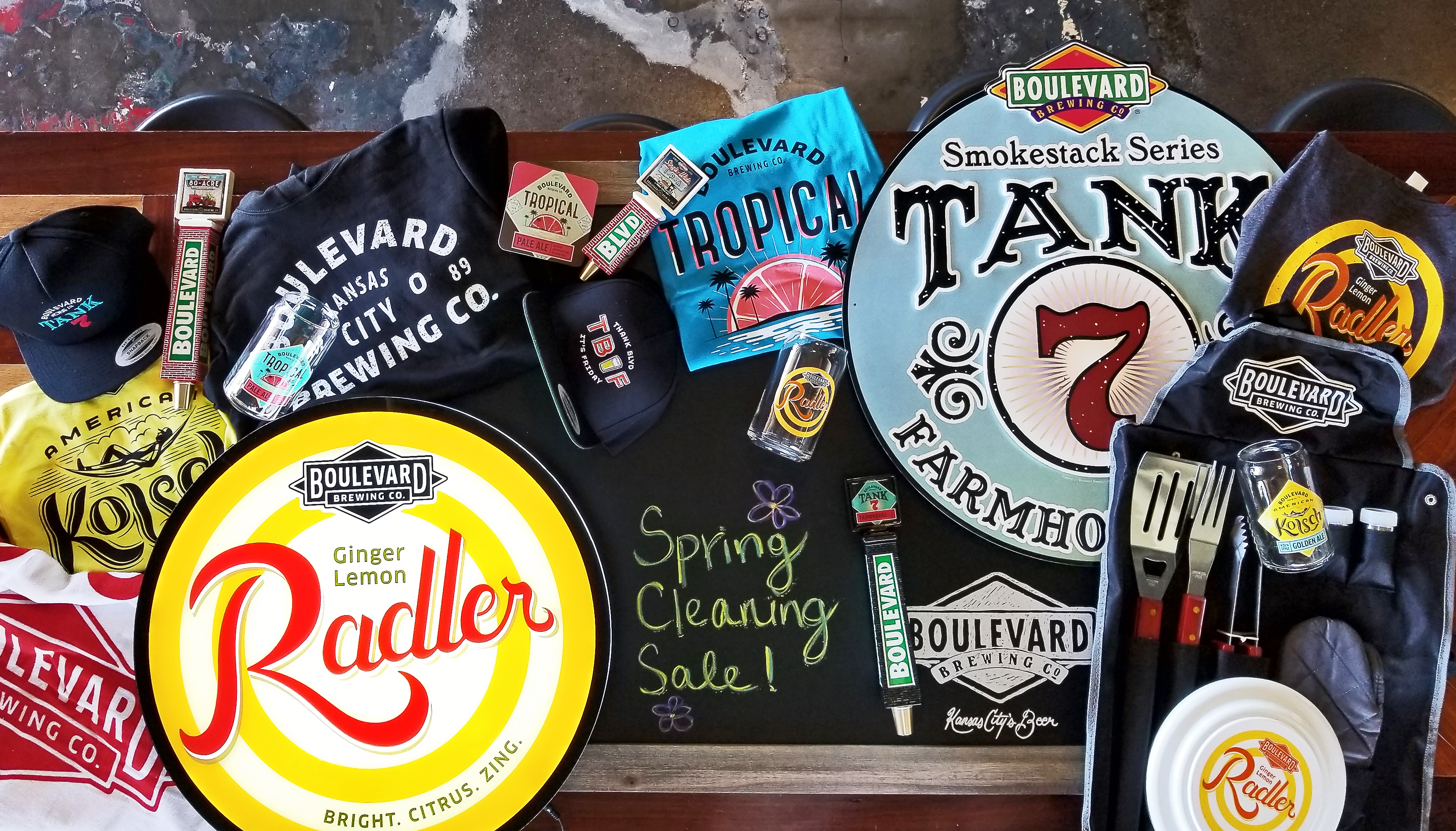 A tabletop displays Boulevard Brewing Company merchandise, including hats, shirts, bottle openers, and coasters. There's a chalkboard with "Spring Cleaning Sale!" written on it, surrounded by various colorful branded items.