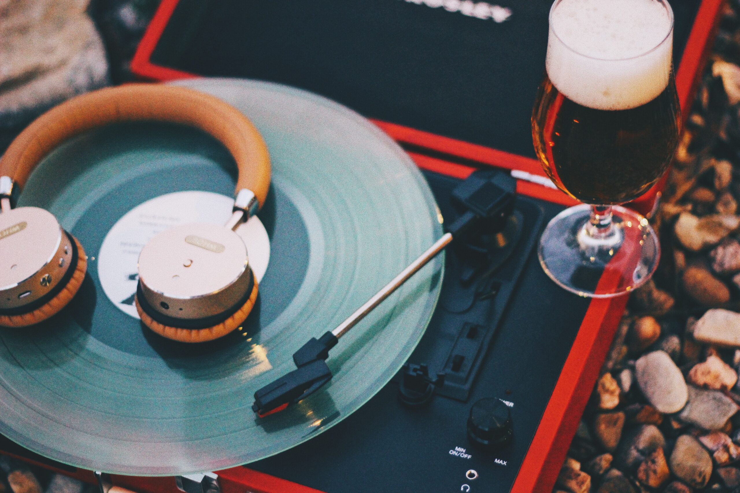 A pair of headphones rests on a spinning record on a red turntable, placed on a bed of pebbles. Beside it, a glass of beer with a frothy head sits on the ground, capturing a relaxed outdoor atmosphere.