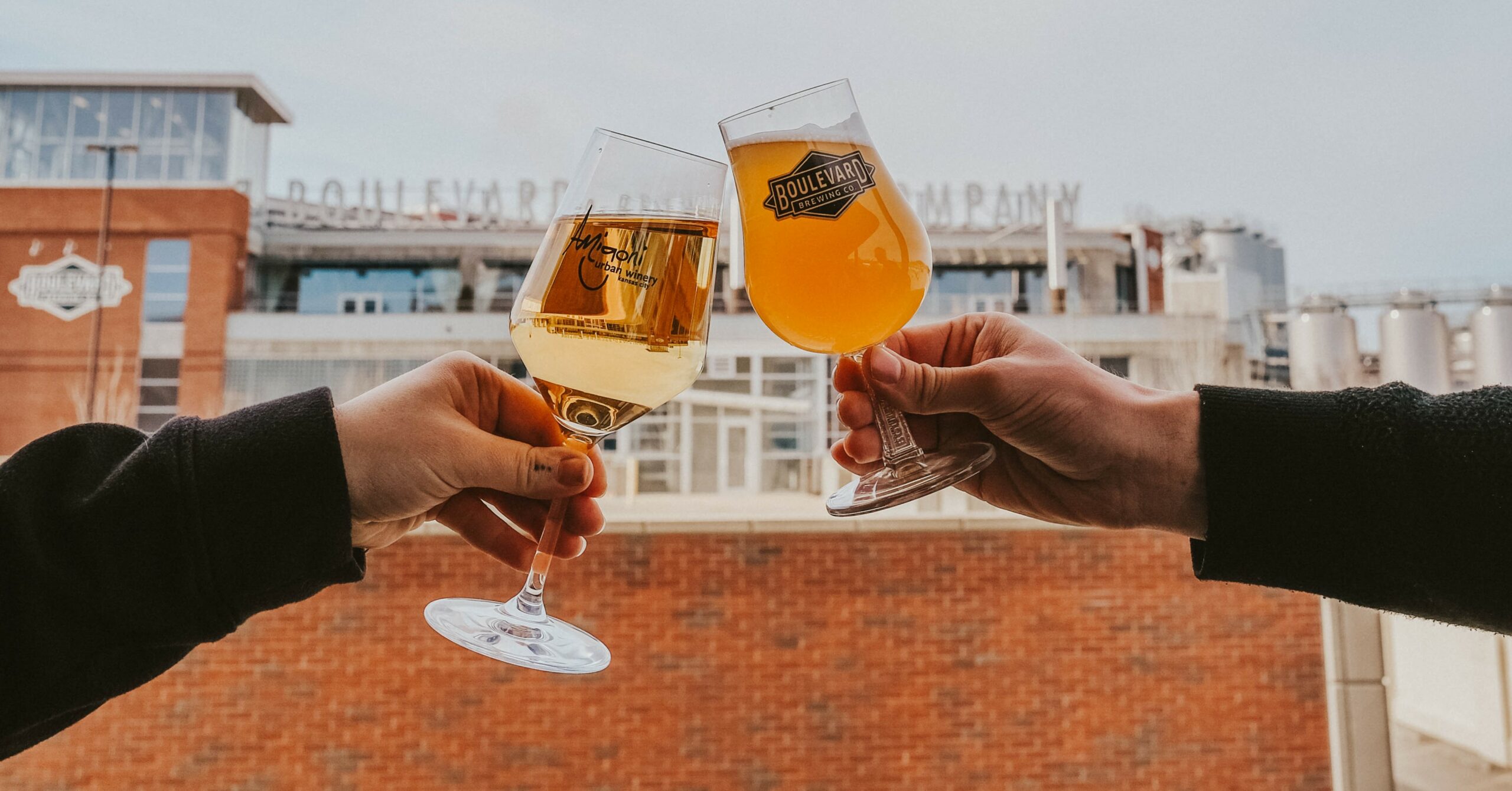 Two people clink glasses of beer in front of Boulevard Brewing Company. One glass contains a lighter-colored beer, while the other holds a darker brew. The sign for the brewing company is visible on the building in the background.
