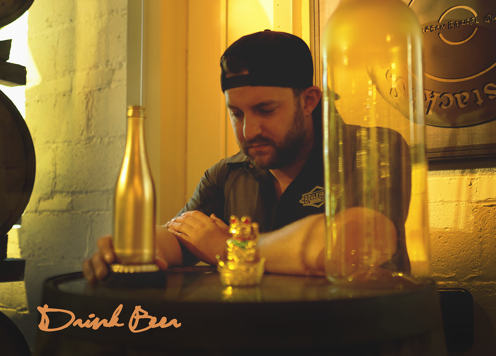 A man wearing a cap is seated at a barrel table with a beer bottle and a gold cat figurine, surrounded by warm lighting. The text "Drink Beer" is written in the bottom left corner.